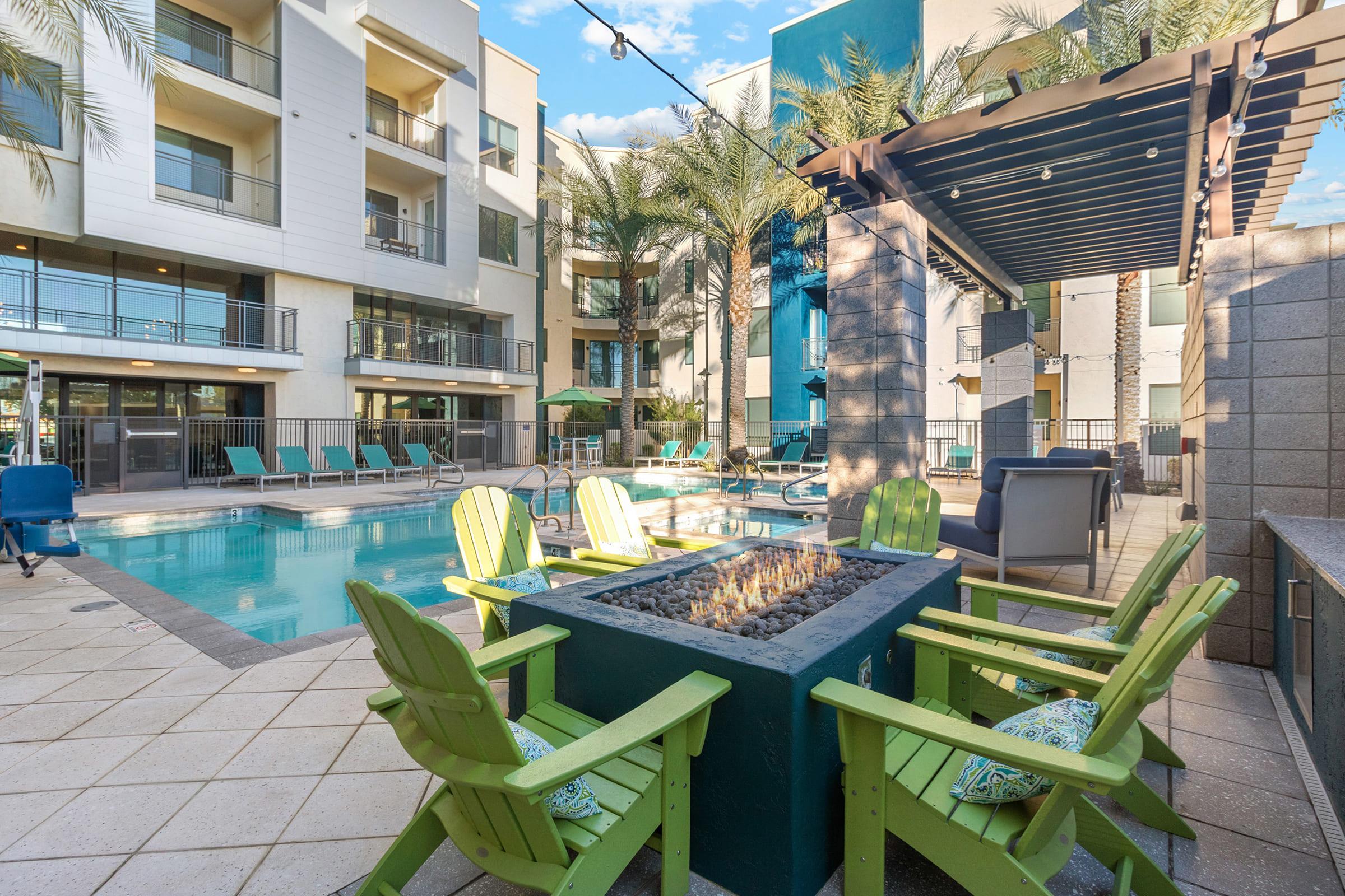 A vibrant outdoor pool area surrounded by modern apartment buildings, featuring a fire pit with green Adirondack chairs, lounge chairs by the pool, and palm trees providing shade. 