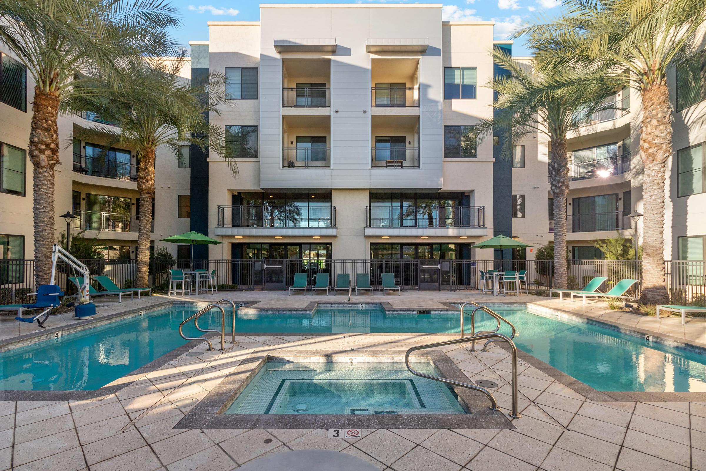 A modern apartment complex with a central swimming pool and hot tub. Surrounding the pool are palm trees and lounge chairs. 