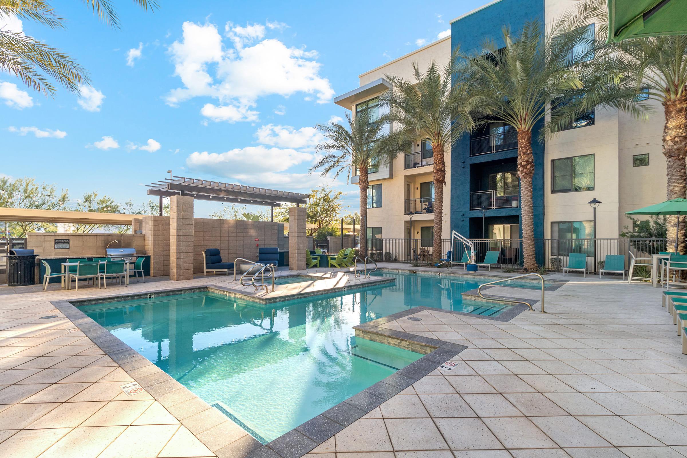 A sparkling swimming pool surrounded by palm trees and lounge chairs, with a modern building in the background. 