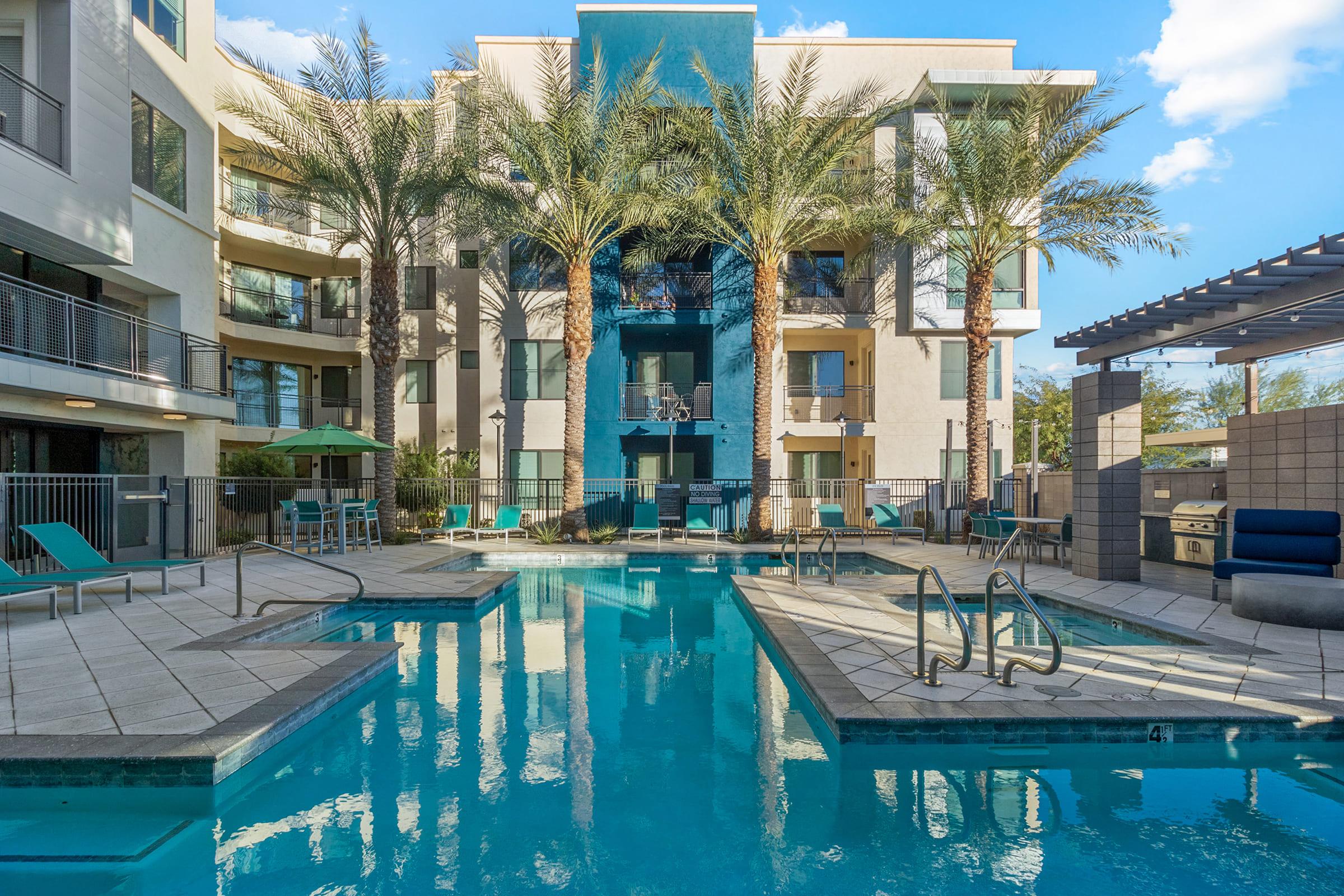 A view of a modern apartment complex featuring a sparkling blue swimming pool surrounded by palm trees.