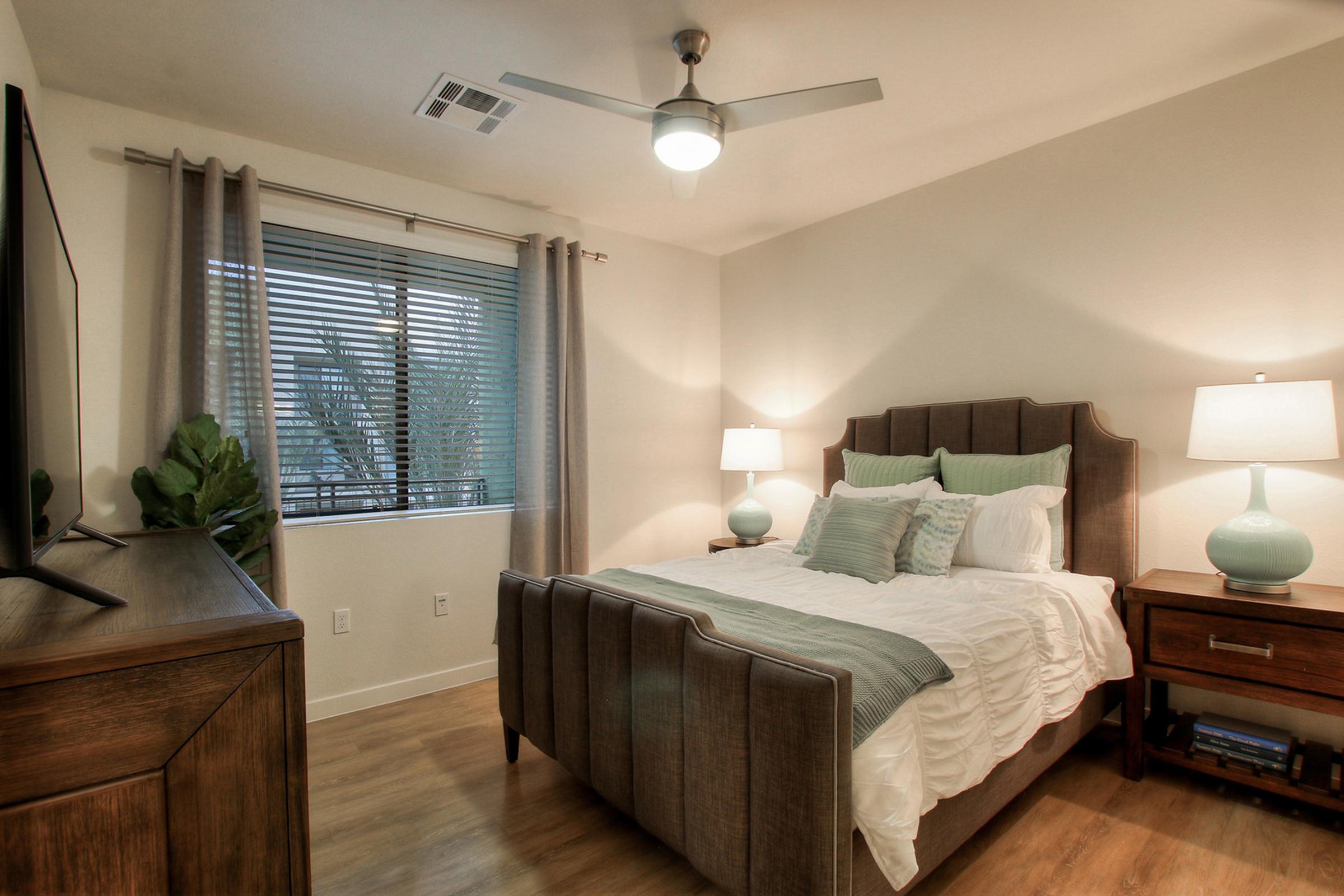  A cozy bedroom featuring a large bed with decorative pillows and a quilt, two bedside lamps on wooden nightstands, a television on a dresser, and a window with light gray curtains partially open, allowing natural light to enter.