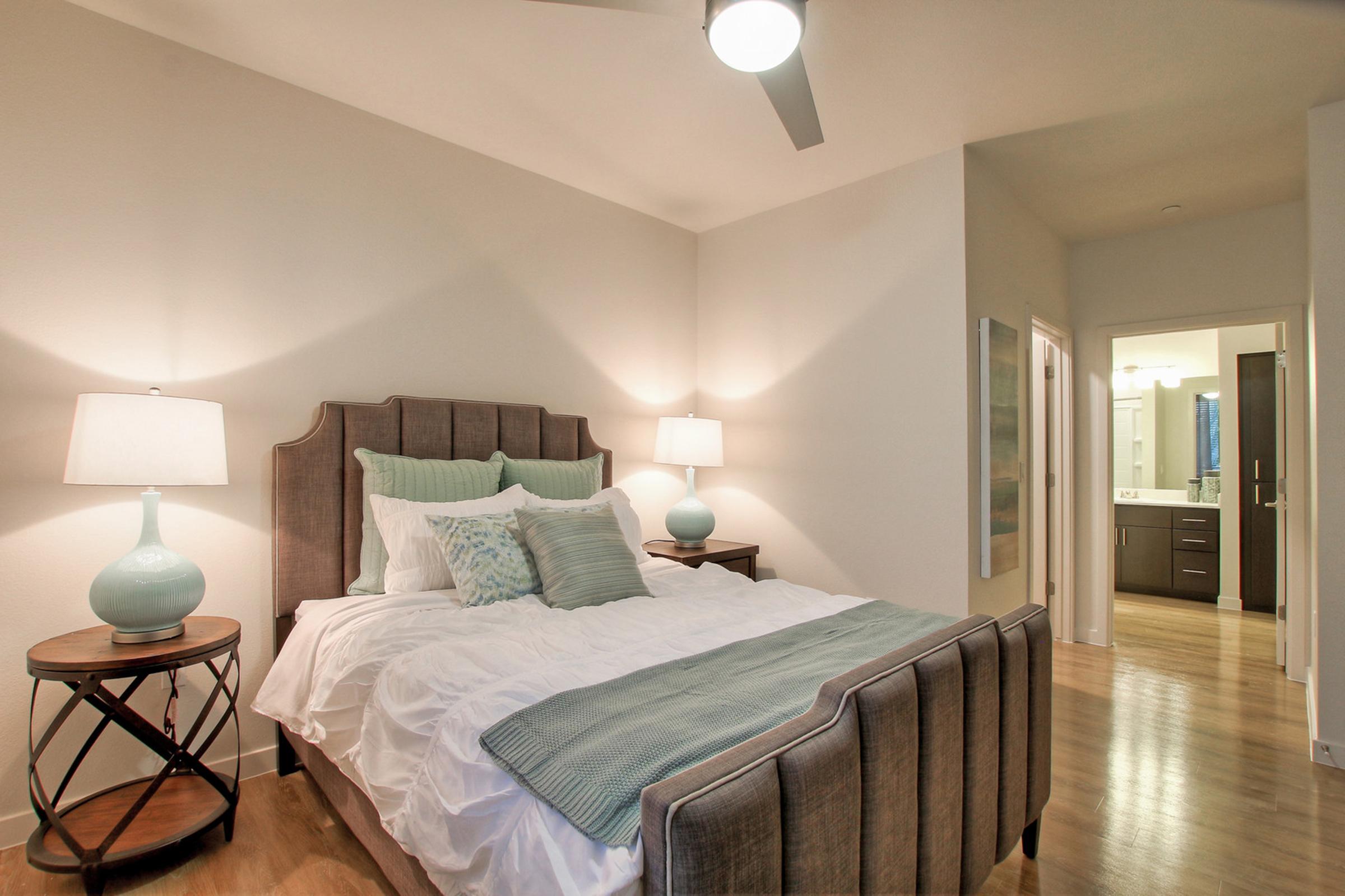 A well-decorated bedroom featuring a queen-sized bed with light blue and white bedding, two bedside lamps on wooden tables, a ceiling fan overhead, and a neutral-colored wall. 