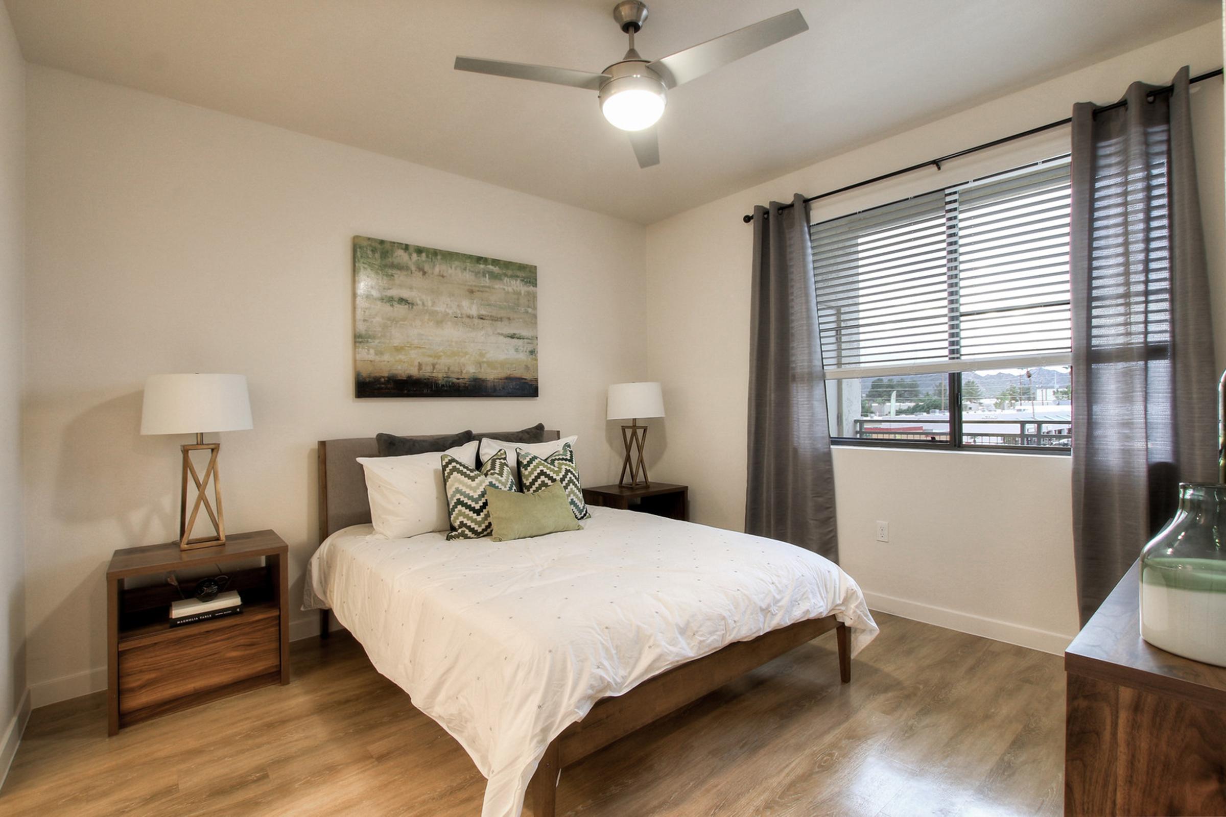 A modern bedroom featuring a double bed with white bedding and decorative pillows, two nightstands with lamps on either side, a large window with blinds, and a piece of abstract art on the wall.