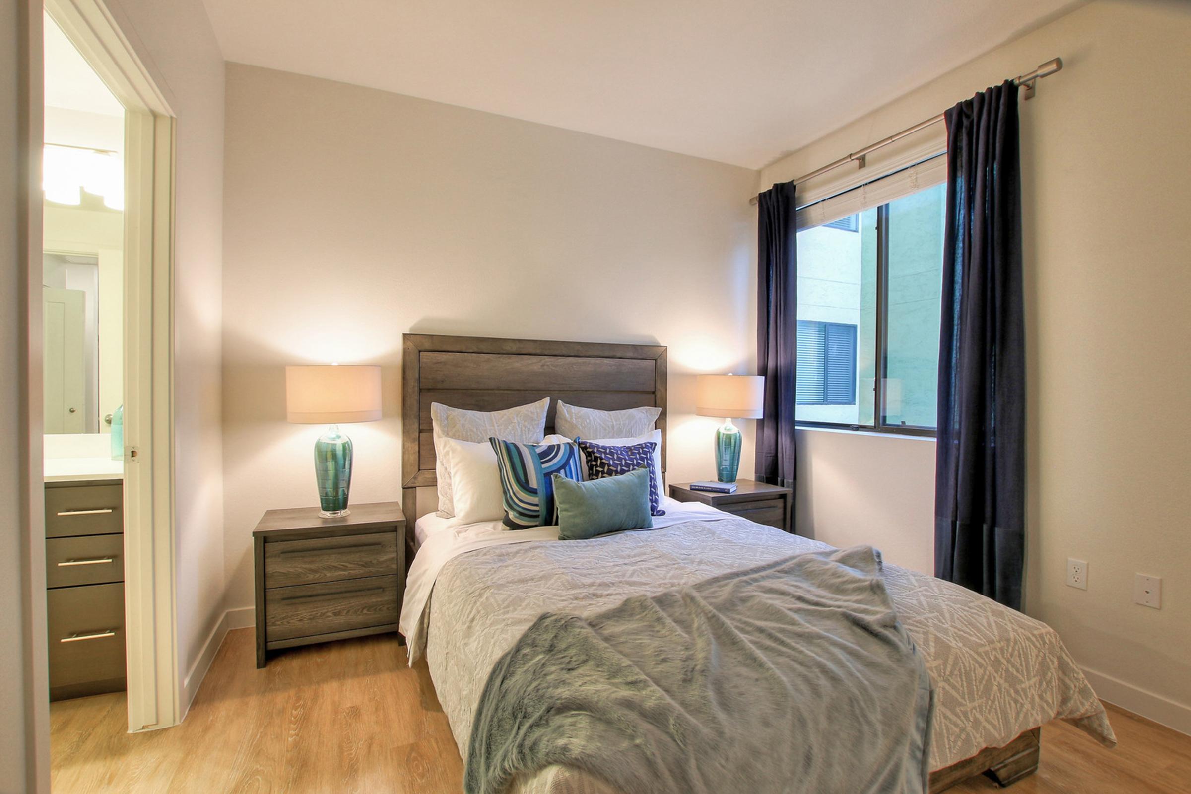  A cozy, well-decorated bedroom featuring a large bed with decorative pillows, two nightstands with blue vases and lamps on either side, and a window adorned with dark curtains.