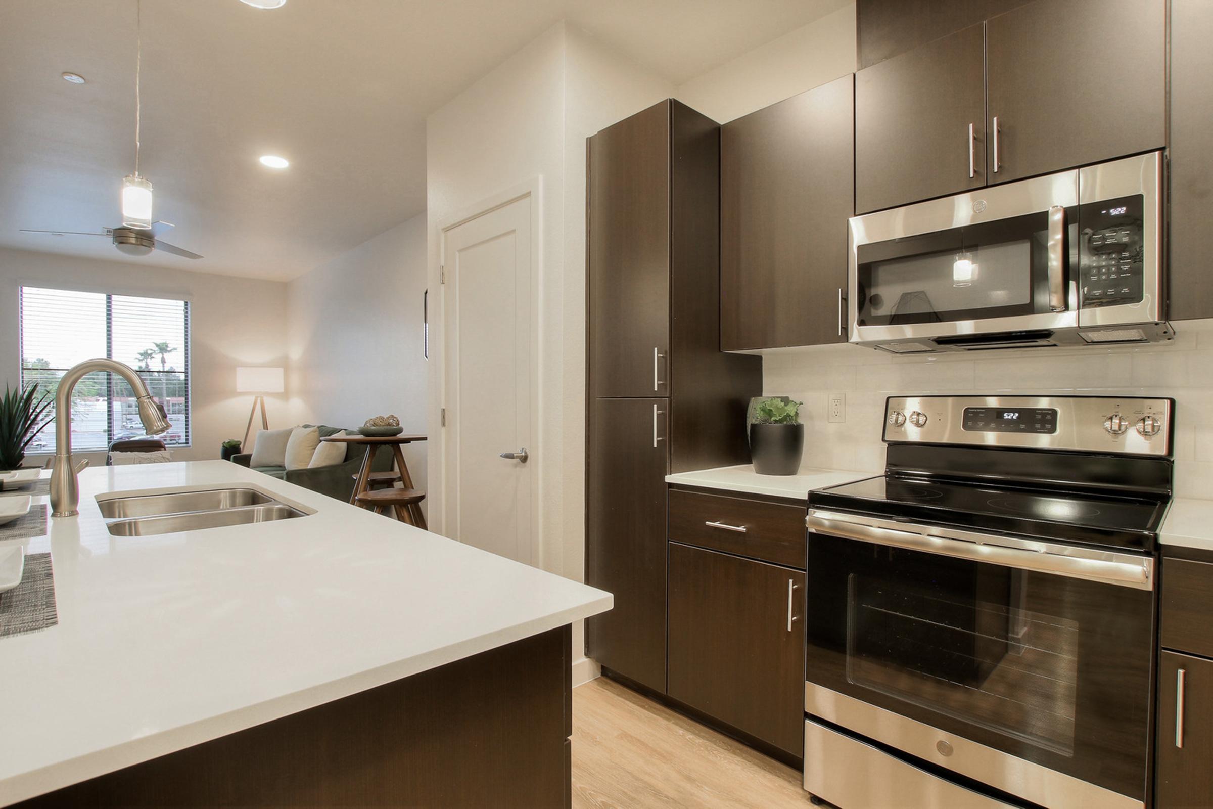  A modern kitchen featuring dark wood cabinetry, a stainless steel oven and microwave, a white countertop, and a sleek sink. 