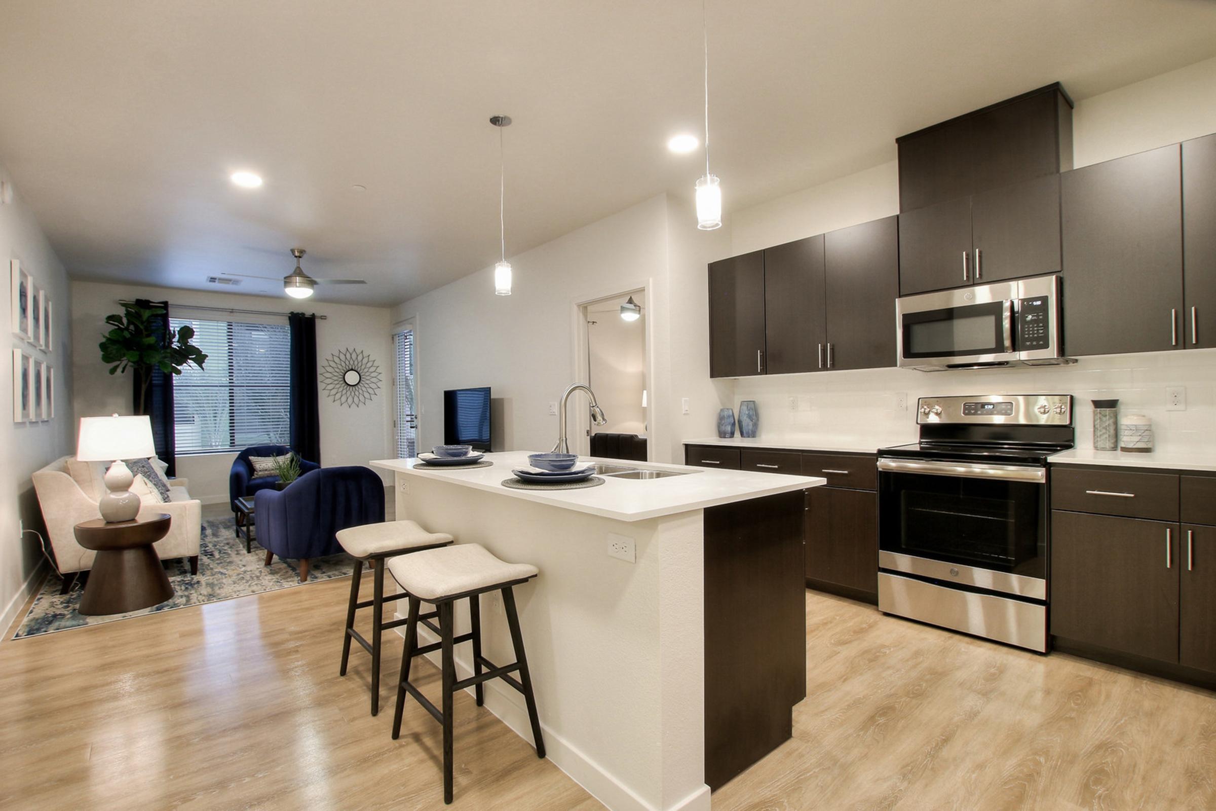 Modern kitchen and living area in an apartment. Features dark wood cabinets, stainless steel appliances, and a kitchen island with two bar stools. 