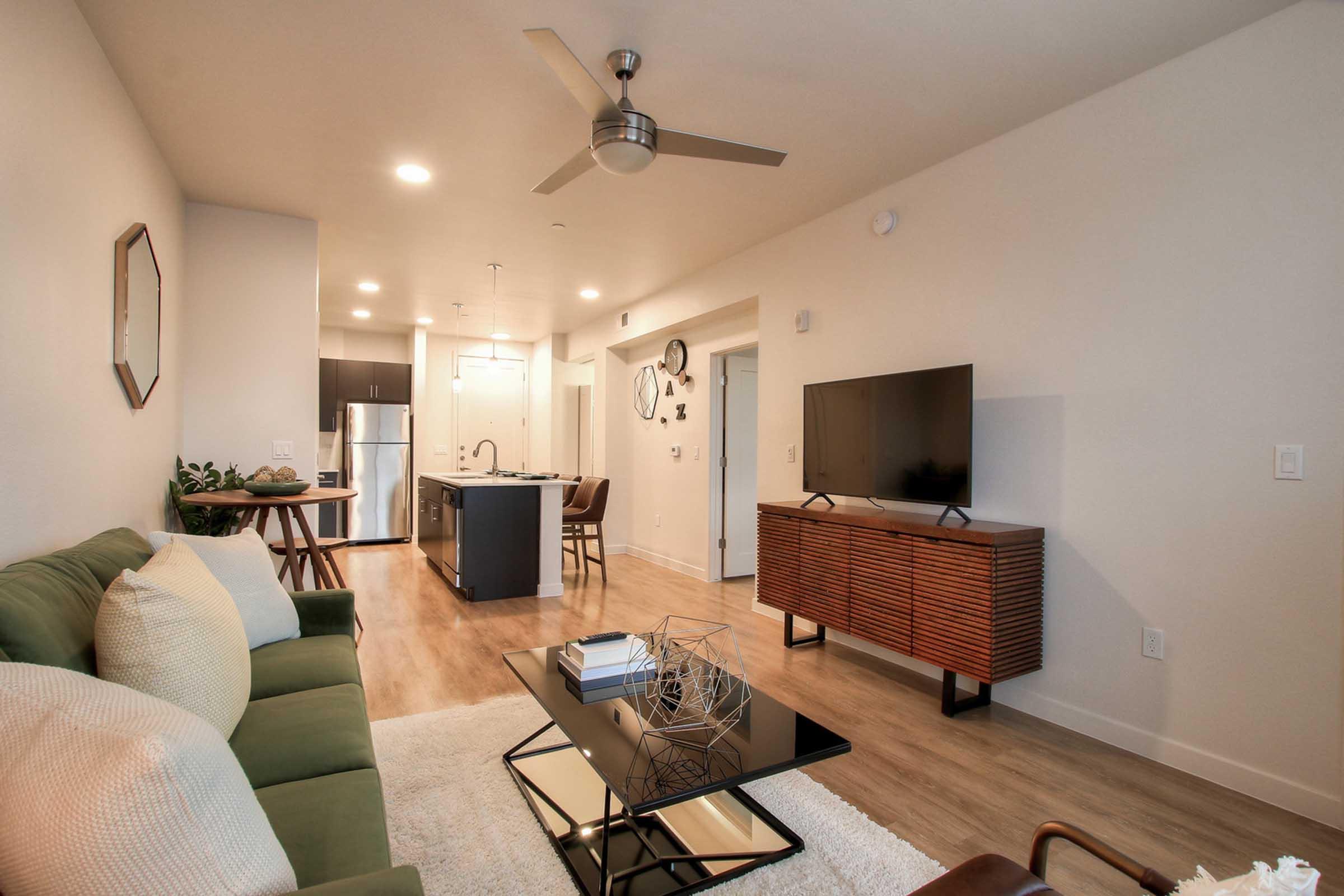 A modern apartment interior featuring a green sofa, a black coffee table with decorative elements, a wooden media console with a TV, and an open kitchen with stainless steel appliances. 