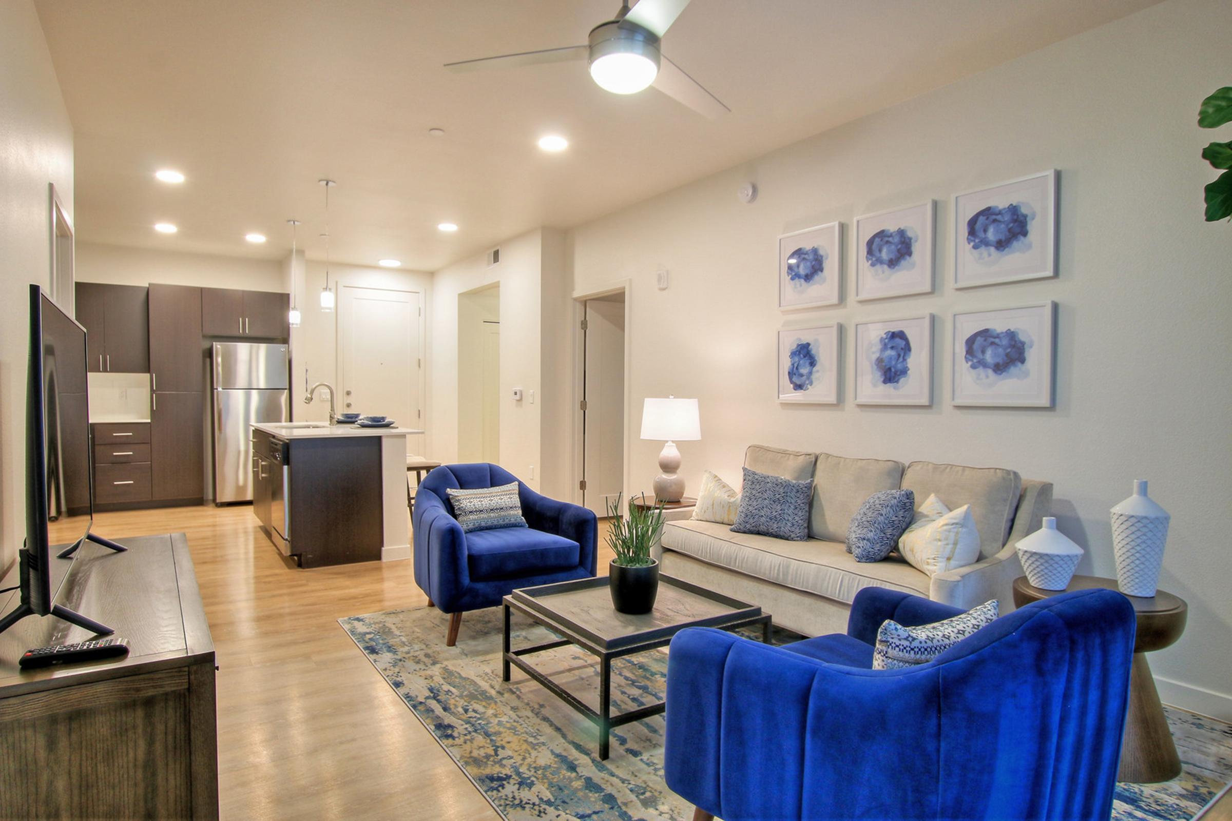 A modern living room featuring a light-colored sofa with blue accent pillows, two vibrant blue chairs, a coffee table, and a TV on a wooden cabinet.