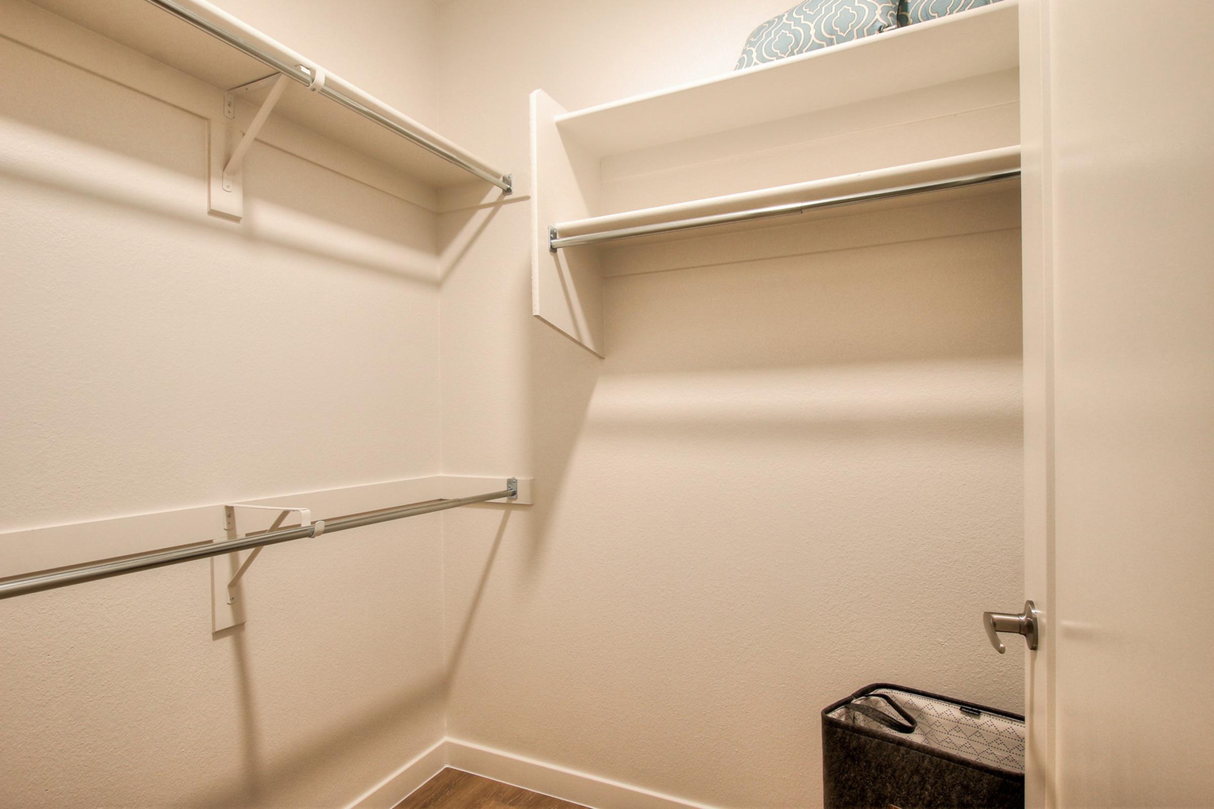 A spacious, empty closet featuring two racks for hanging clothes, a shelf above, and a small laundry basket in the corner. 