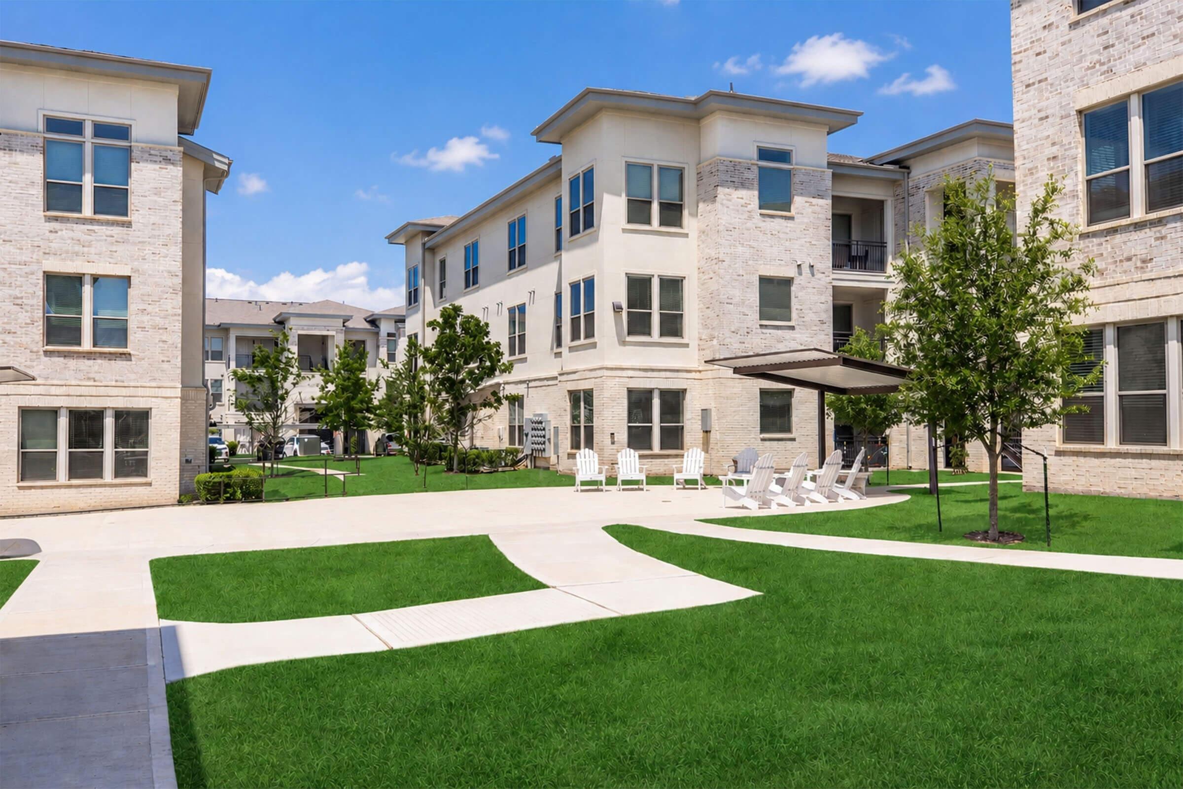Modern apartment complex with light-colored stone exterior, featuring multiple stories. A landscaped courtyard with well-maintained grass, shaded seating areas, and white lounge chairs under a canopy. Clear blue skies in the background complete the inviting residential atmosphere.