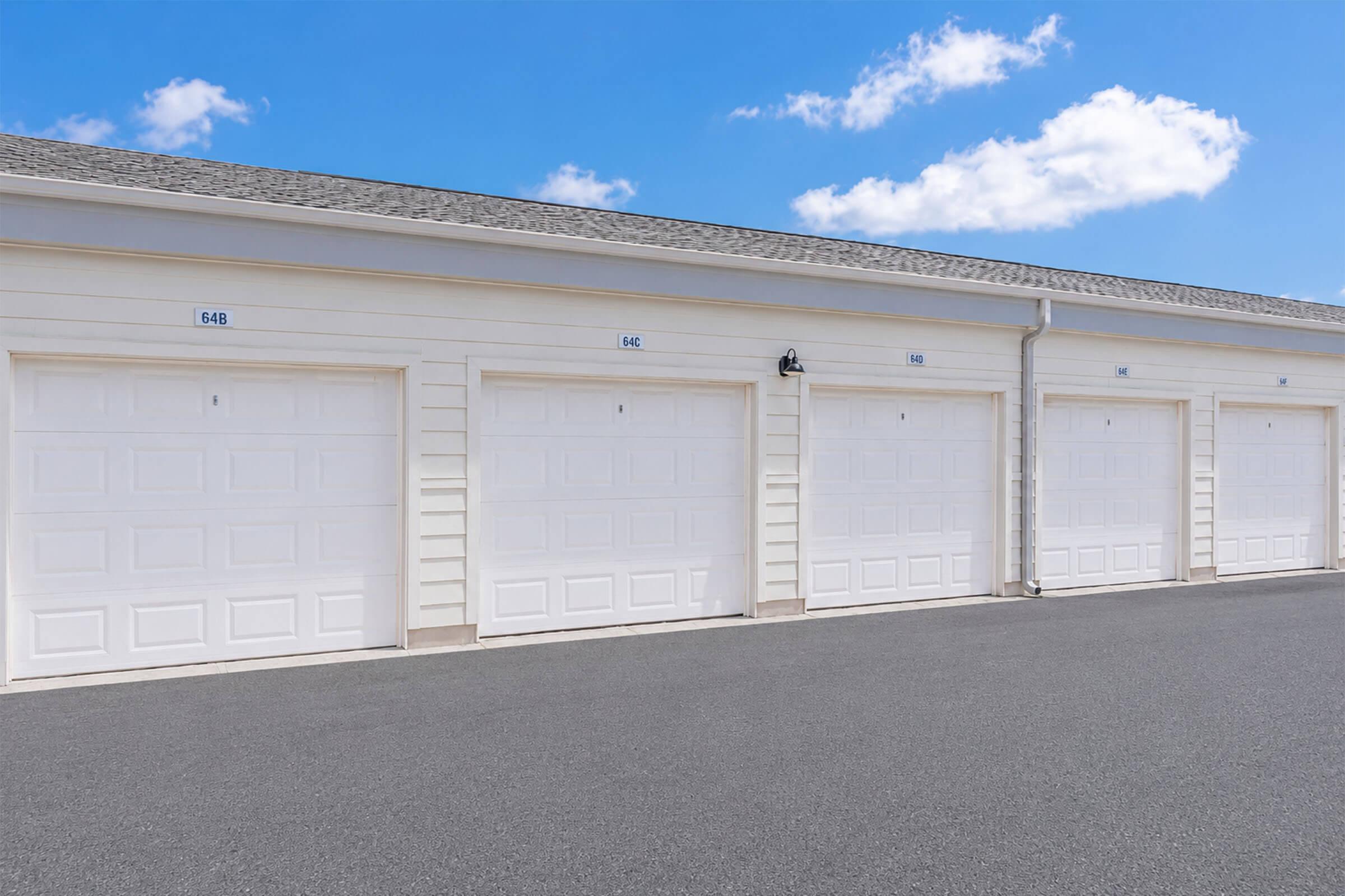 A row of white garage doors on a light-colored building, with blue sky and clouds in the background. The garage doors are numbered, and there is a clear paved surface in front. The setting appears clean and well-maintained.