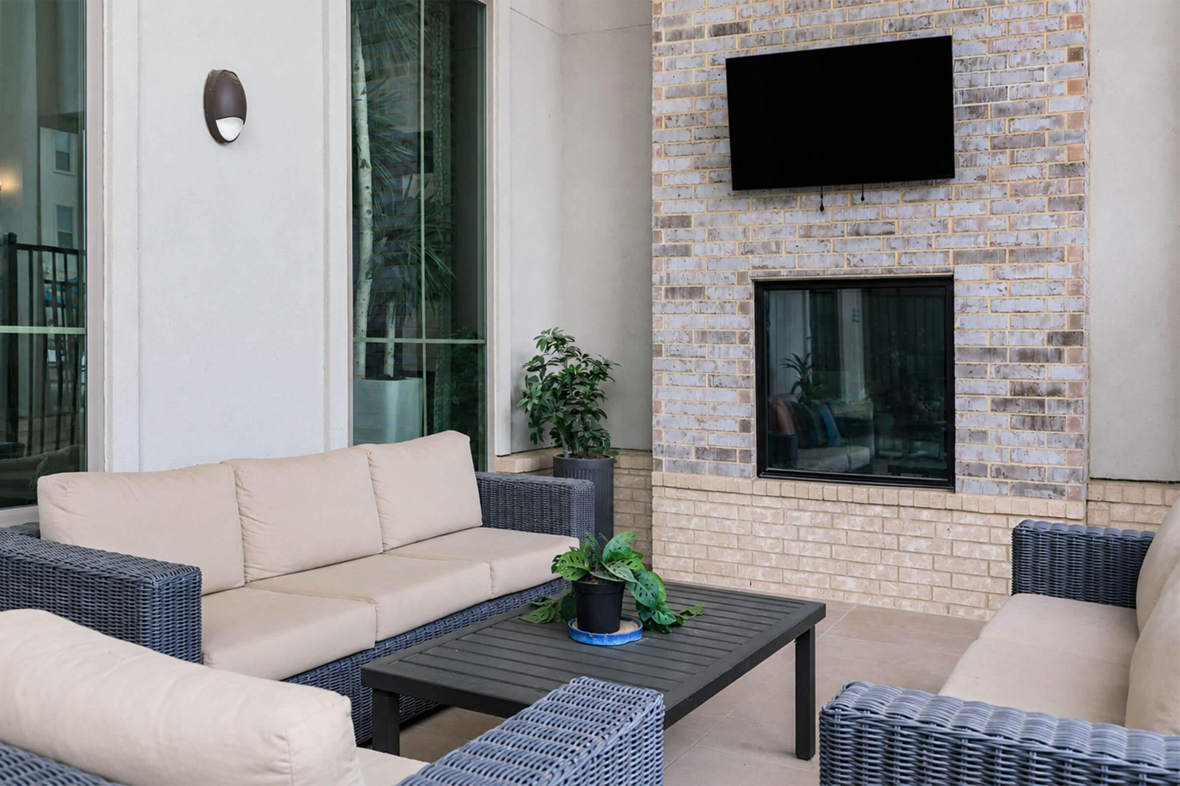 A modern outdoor seating area featuring two beige sofas and a coffee table on a patio. The wall behind has a decorative stone fireplace and a flat-screen TV mounted above it. Potted plants are present, contributing to a relaxed atmosphere.
