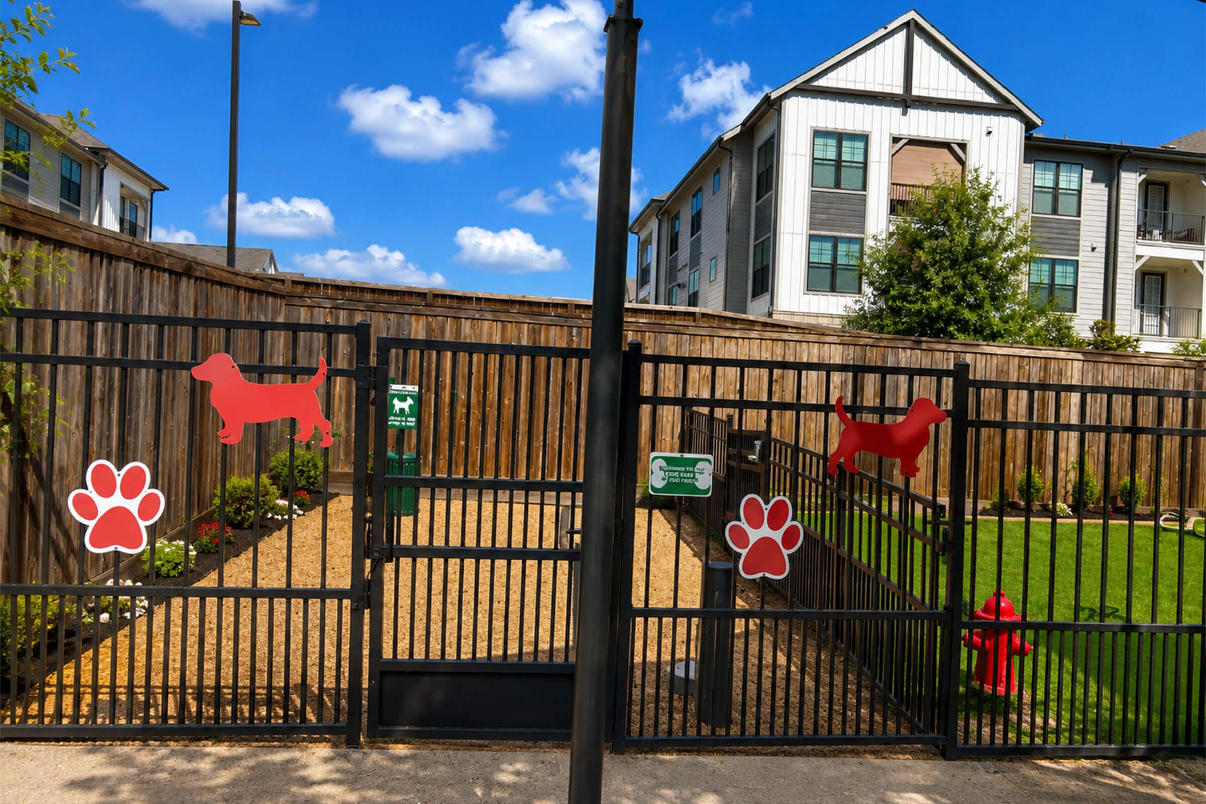 A fenced dog park with red paw print decorations. Inside, there are grassy areas and a dog-friendly fire hydrant. The background features an apartment building with a mix of wooden and modern elements under a blue sky with fluffy clouds.