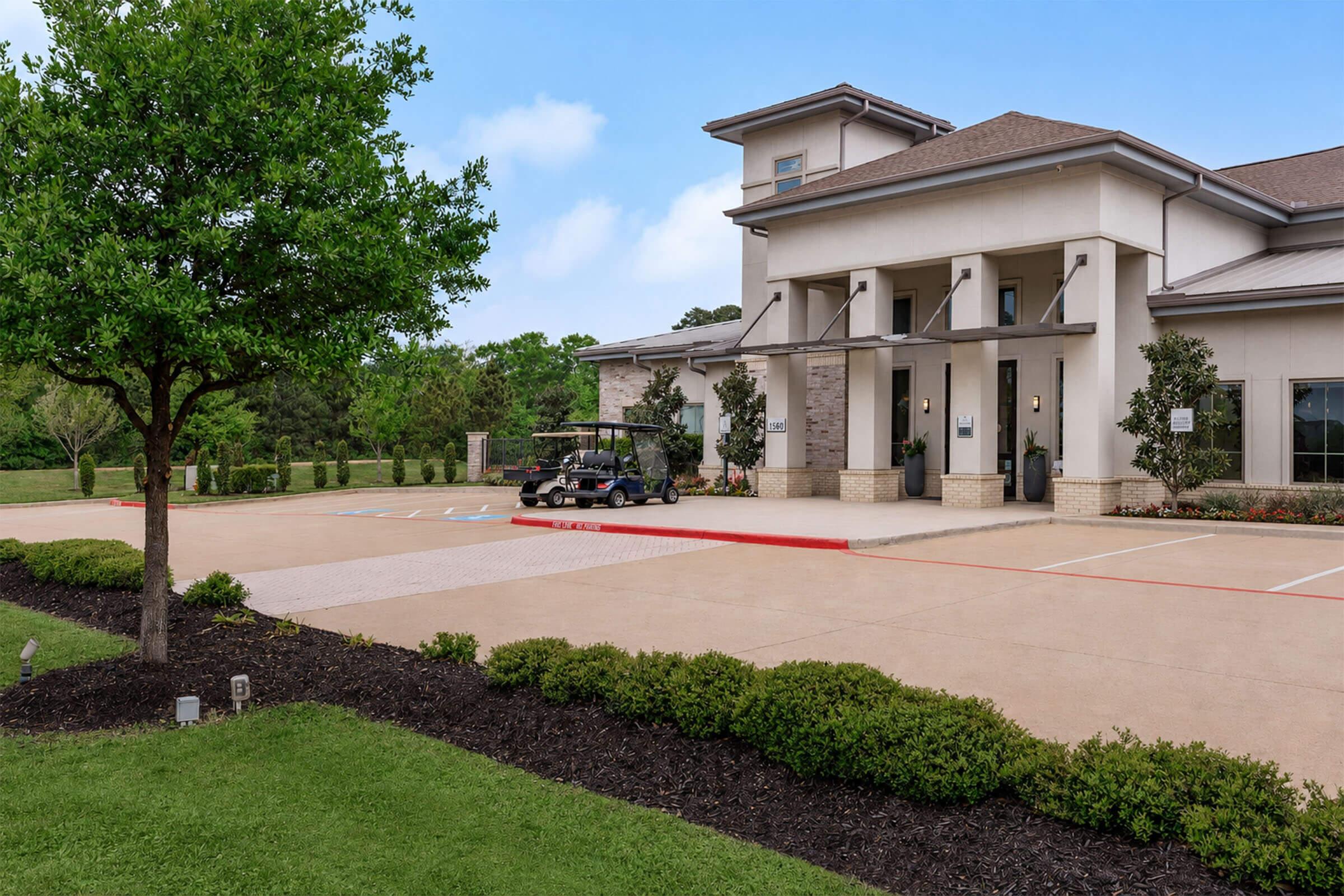 A modern building with large entrance doors, flanked by neatly landscaped trees and shrubs. In the foreground, there is a well-maintained parking area with a golf cart parked. The surroundings are green and inviting, with a clear blue sky above.