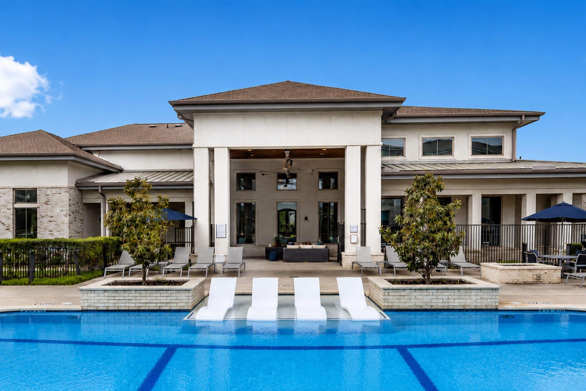 Modern building with a patio overlooking a blue swimming pool. The pool features white lounge chairs and umbrellas. Lush greenery borders the area, and the sky is clear with a few clouds. The architecture is contemporary, showcasing large windows and an inviting outdoor space.