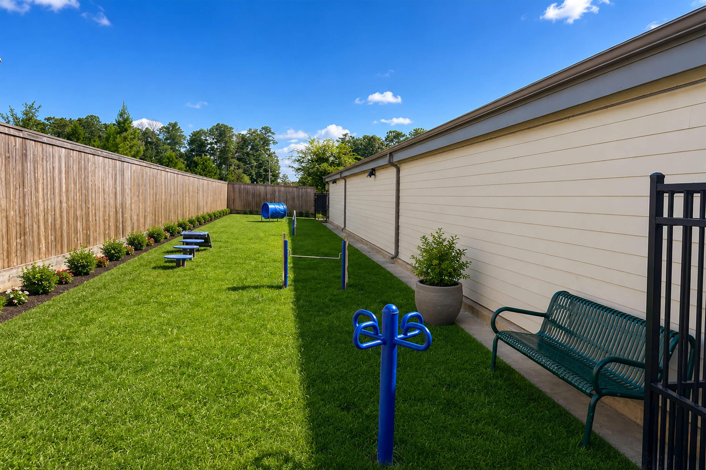 A landscaped outdoor area featuring a manicured lawn, exercise equipment in blue, and a green bench. There are planters with plants along the sides, and a wooden fence in the background. The sky is clear with a few clouds, creating a bright and inviting atmosphere.