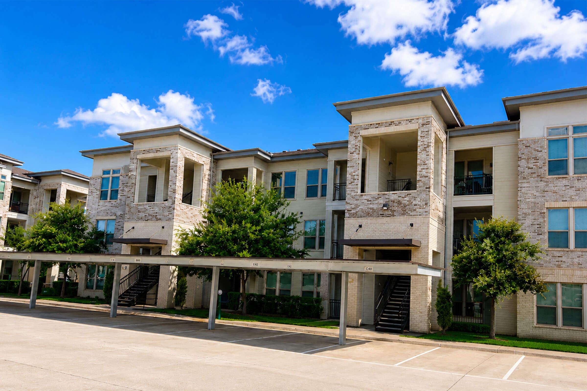 A modern apartment building featuring multiple stories with stone and siding exteriors. The structure has balconies, greenery around the base, and covered parking spaces in the foreground. The sky is bright blue with fluffy white clouds, creating a welcoming atmosphere.