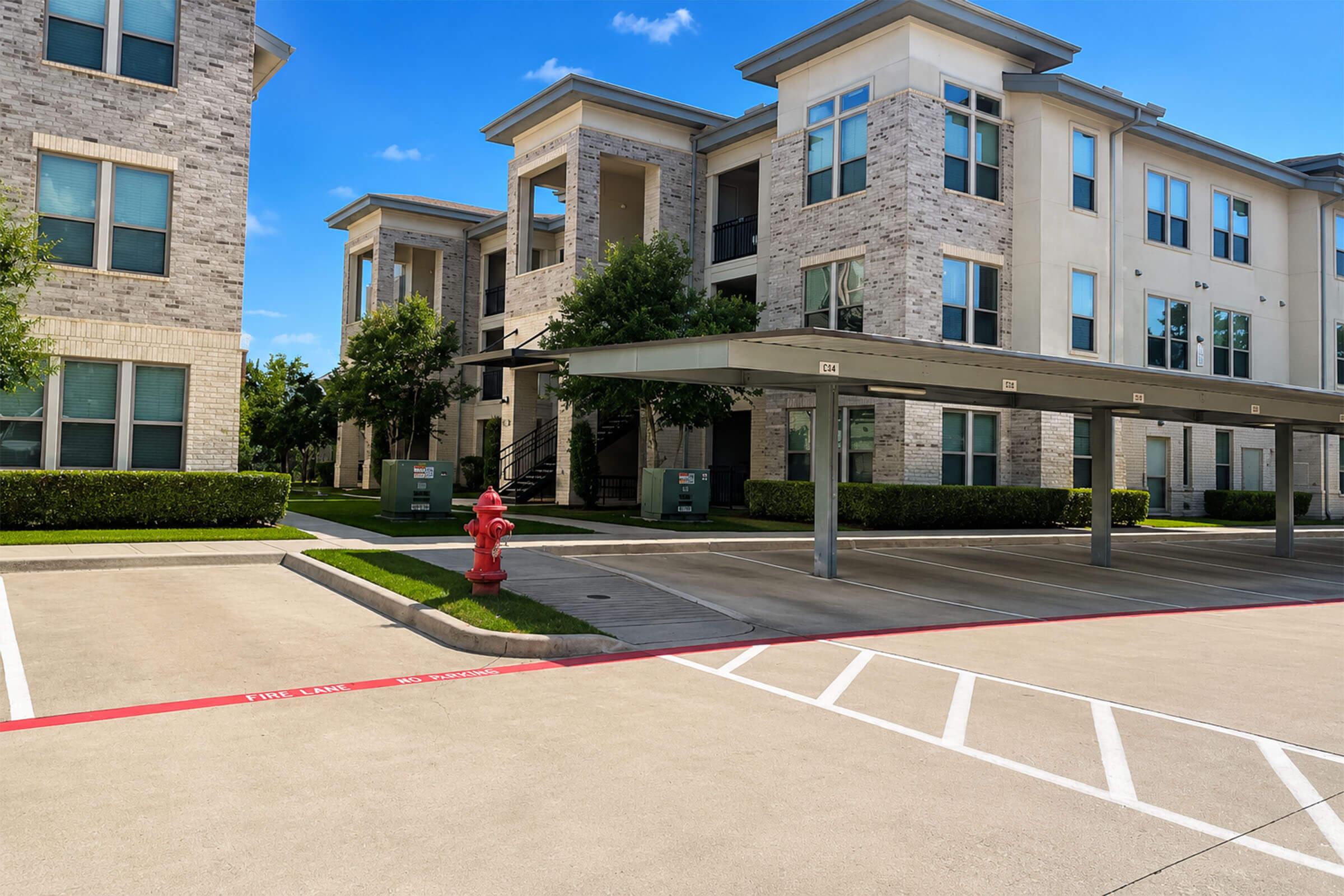 A residential apartment complex featuring multiple buildings with modern architecture. There is a covered parking area and a red fire hydrant near the driveway, surrounded by well-maintained landscaping and green grass under a clear blue sky.