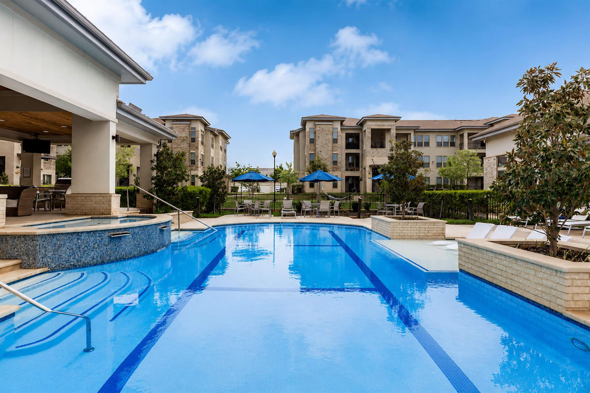 A serene swimming pool with clear blue water, surrounded by a modern apartment complex. There are lounge chairs and blue umbrellas near the pool area, with green landscaping in the background and a bright blue sky above. The scene conveys a relaxing and inviting atmosphere.