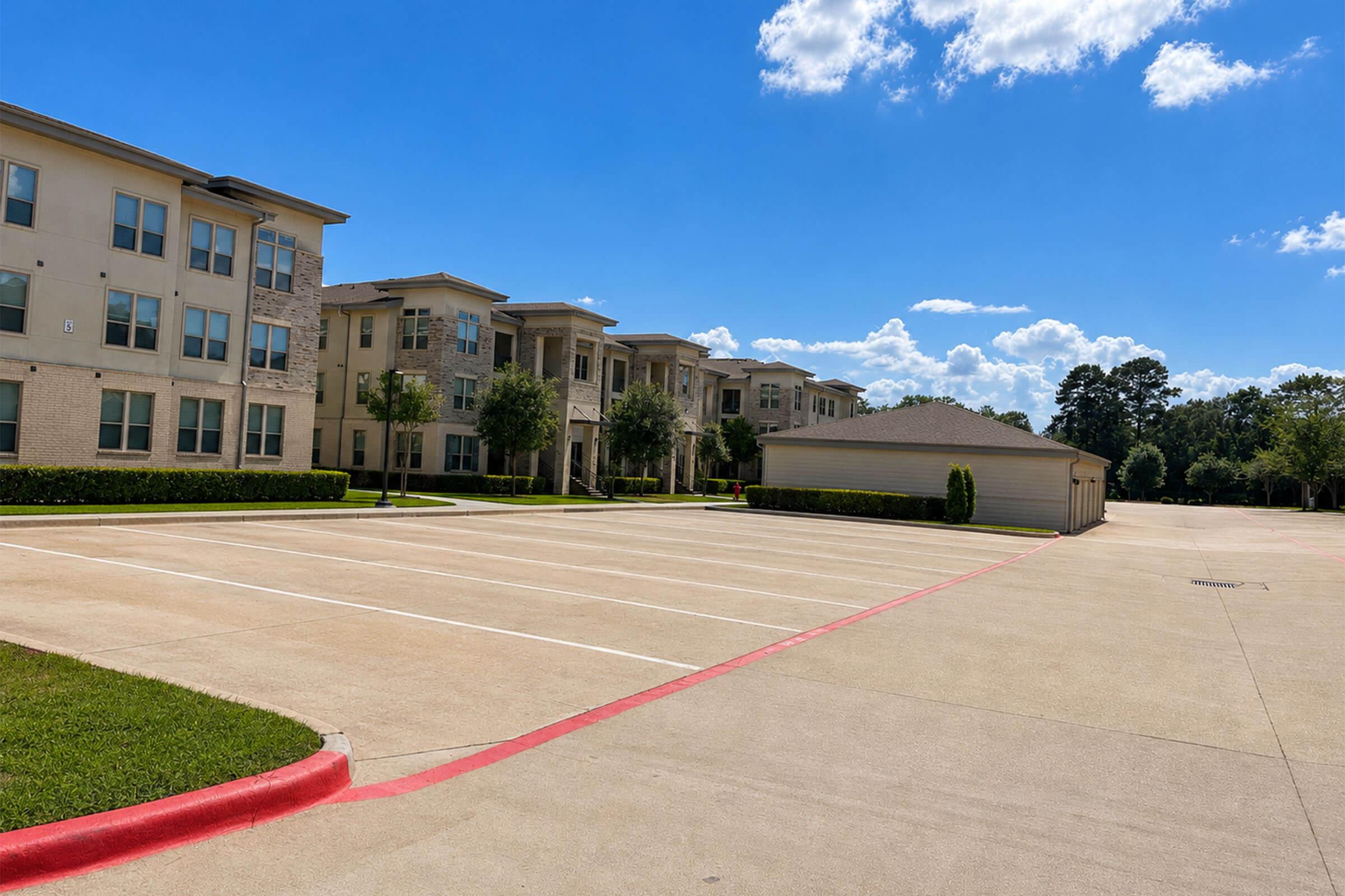 A clear view of a modern apartment complex featuring several buildings with balconies. An empty parking lot is in the foreground, lined with painted white stripes, and a maintenance or storage building can be seen to the right. The sky is bright blue with a few clouds, and there are trees and shrubs around the area.