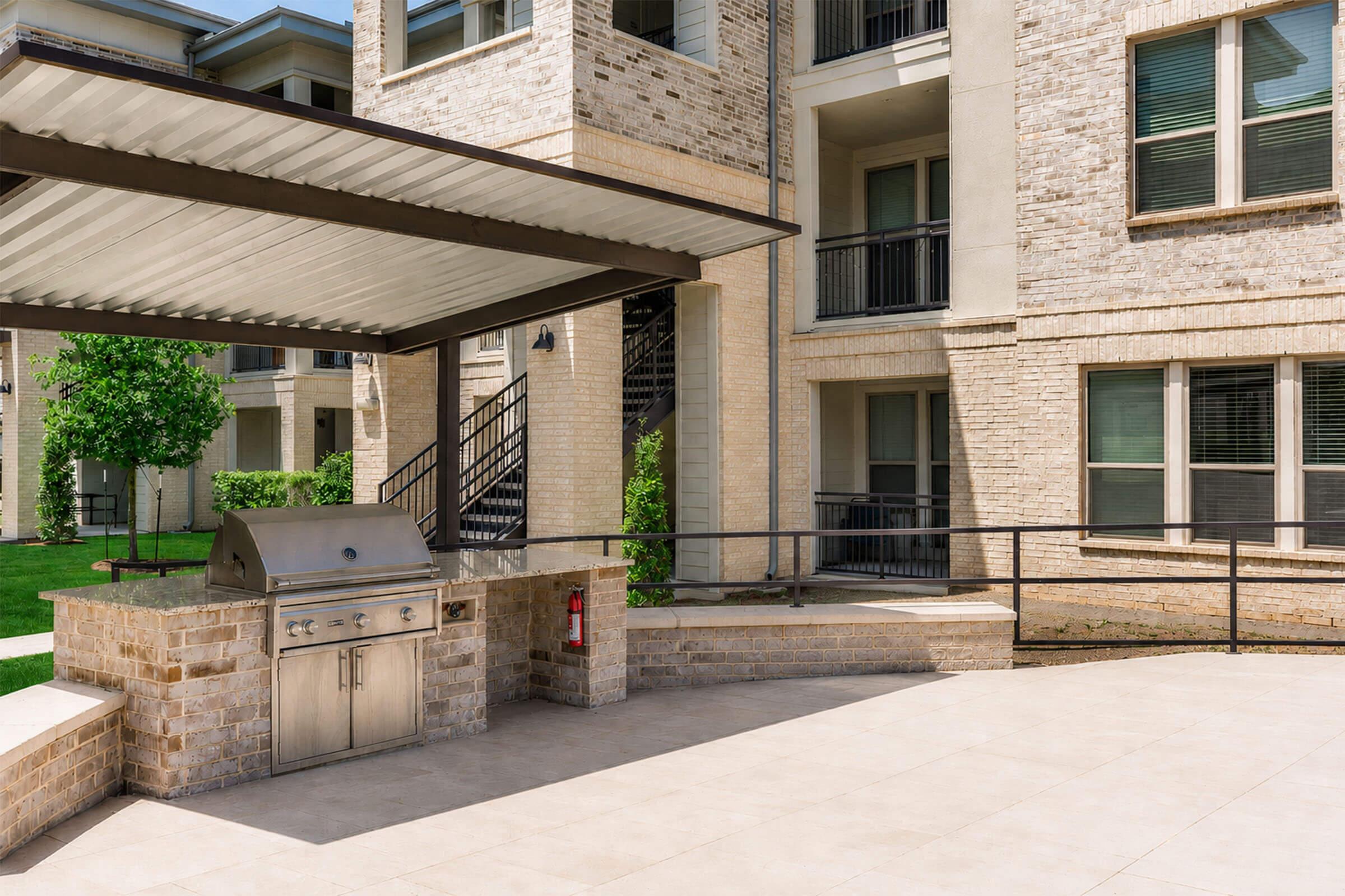 An outdoor barbecue area featuring a stainless steel grill under a shaded pavilion, surrounded by a landscaped courtyard with modern apartment buildings in the background. The setting includes stone and concrete elements, with green trees adding a pop of color.