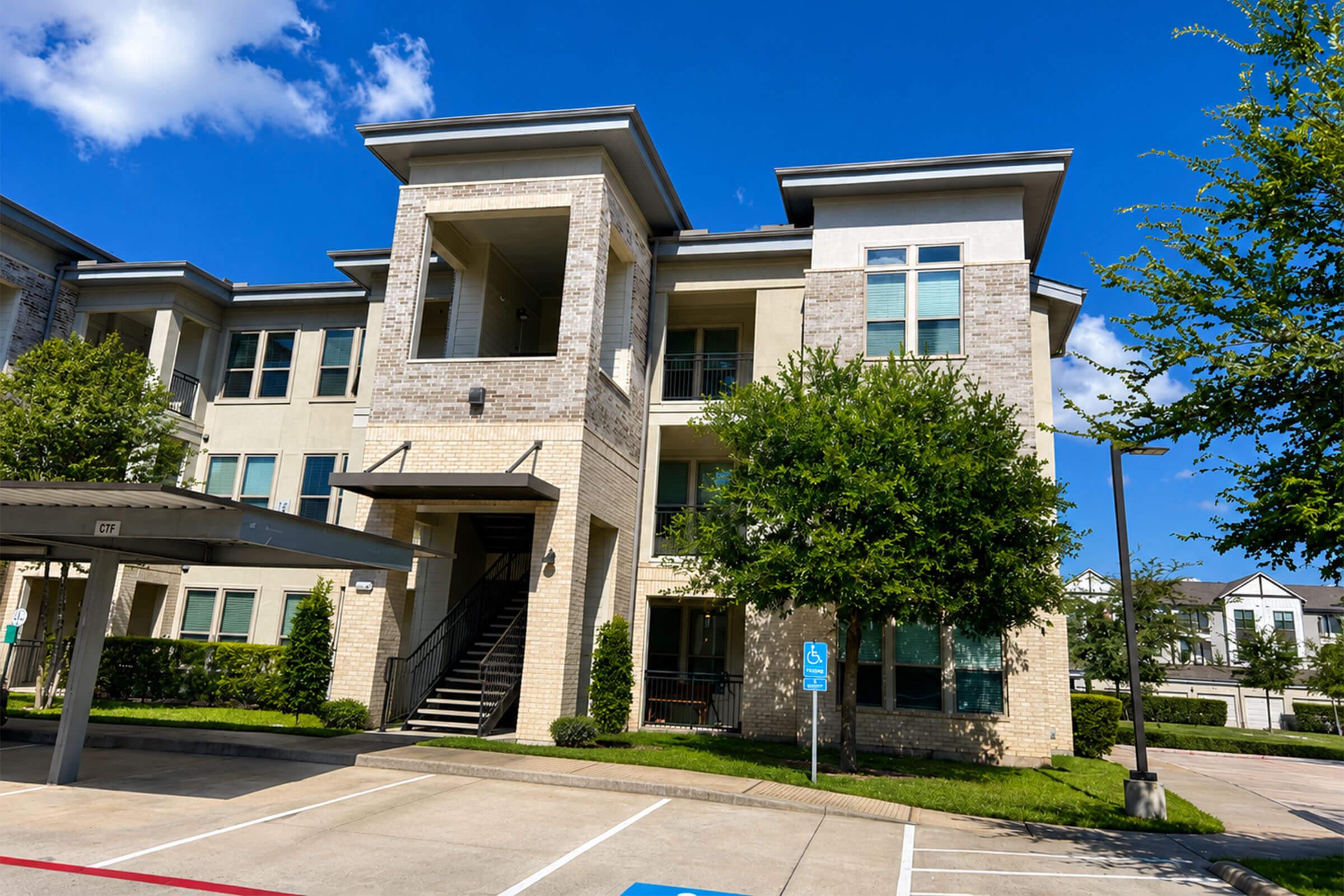 Modern apartment building with a stone and stucco exterior, featuring multiple balconies and large windows. Surrounding greenery includes small trees and shrubs. The sky is clear and blue, with a few clouds, and there are parking spaces visible in the foreground.