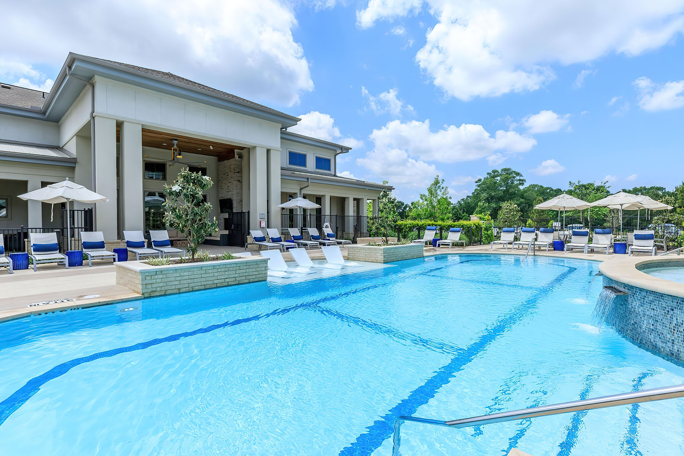 A luxurious outdoor swimming pool surrounded by lounge chairs and umbrellas, adjacent to a modern building. The pool features clear blue water, with a hot tub on one side. Lush greenery and blue skies with fluffy clouds create a relaxing atmosphere. Perfect for leisure and relaxation.