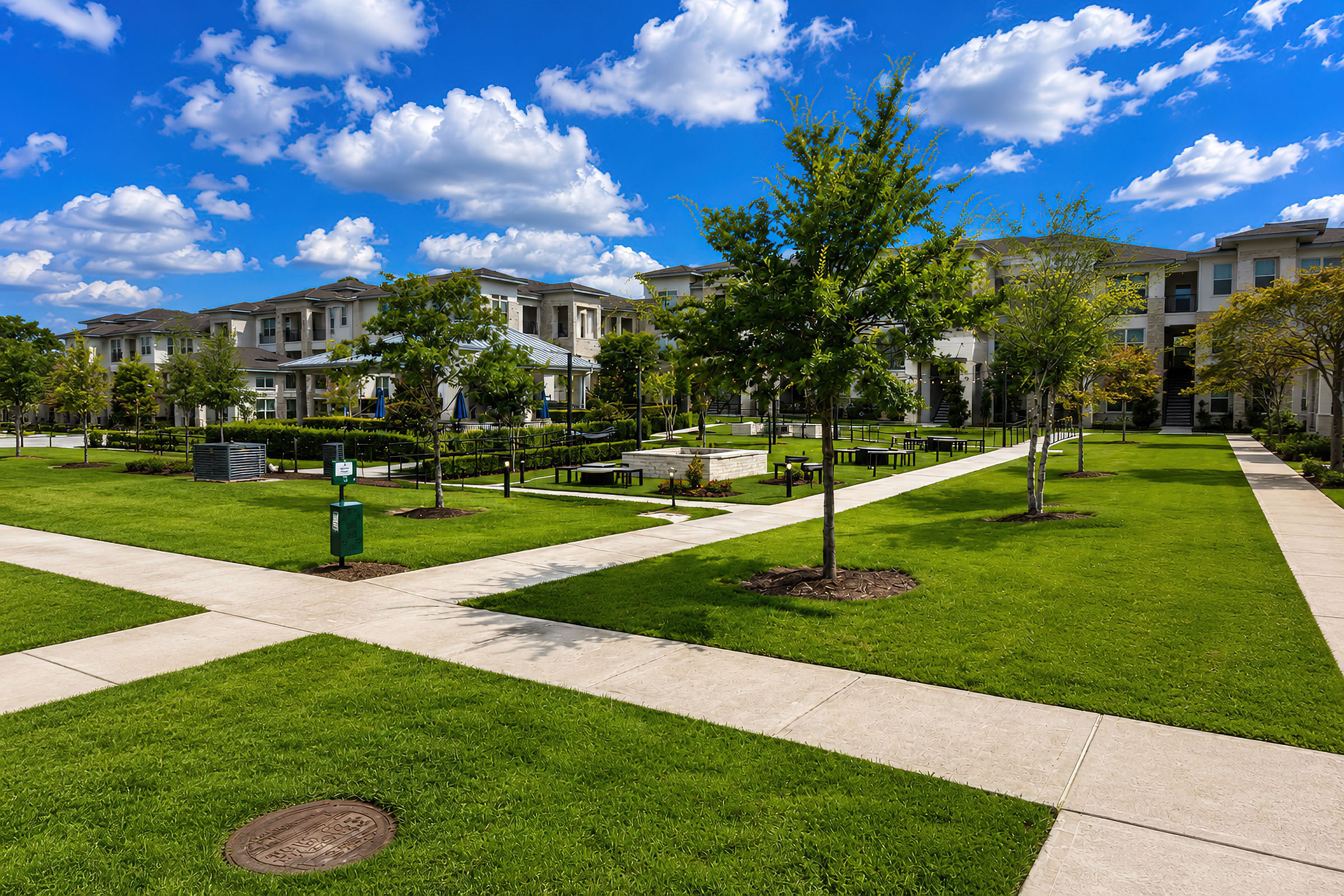 A landscaped courtyard featuring green grass, walking paths, and trees, surrounded by modern apartment buildings. The scene is set under a bright blue sky with fluffy white clouds, creating a serene and inviting atmosphere.