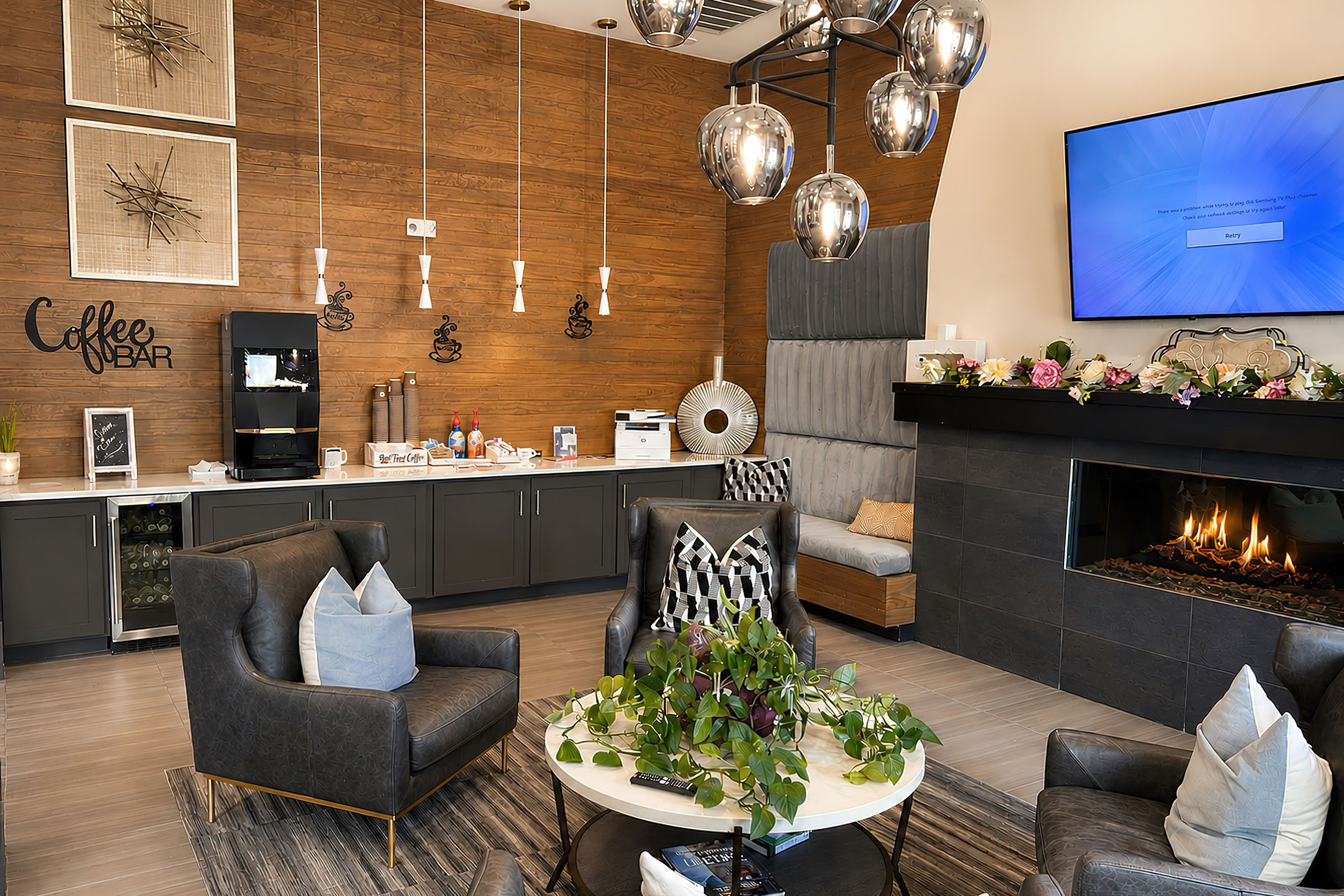 A cozy coffee bar area with modern décor featuring a fireplace, comfortable seating, and pendant lighting. There are plants and decorative elements on a coffee table, along with a coffee machine, snacks, and a TV mounted on the wall, creating an inviting space for relaxation and socializing.
