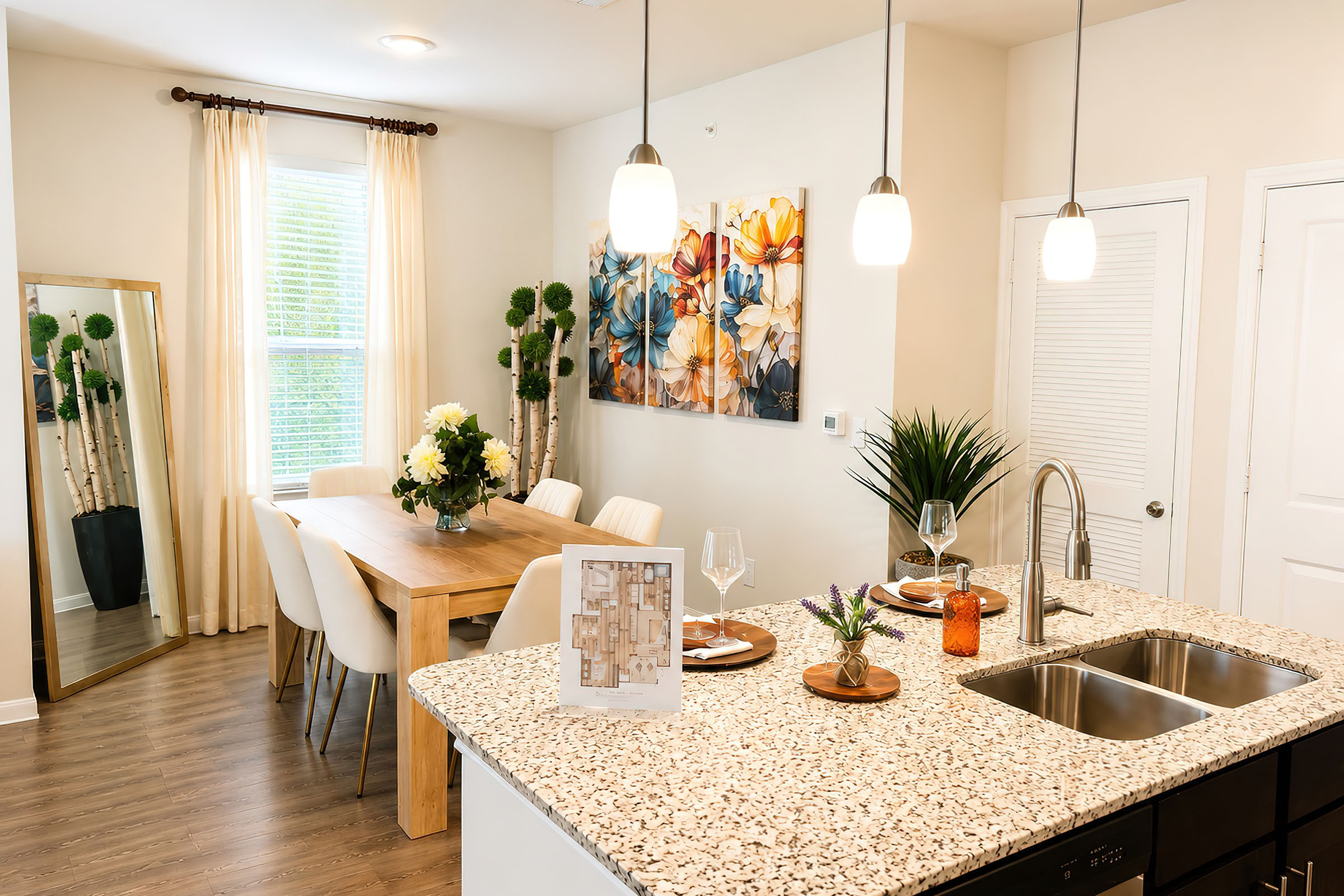 Modern kitchen and dining area featuring a large island with a granite countertop, a dining table set for six with a floral centerpiece, and pendant lighting. The walls are adorned with flower artwork, and there is a large mirror reflecting the room's interior. Natural light flows through a window with sheer curtains.