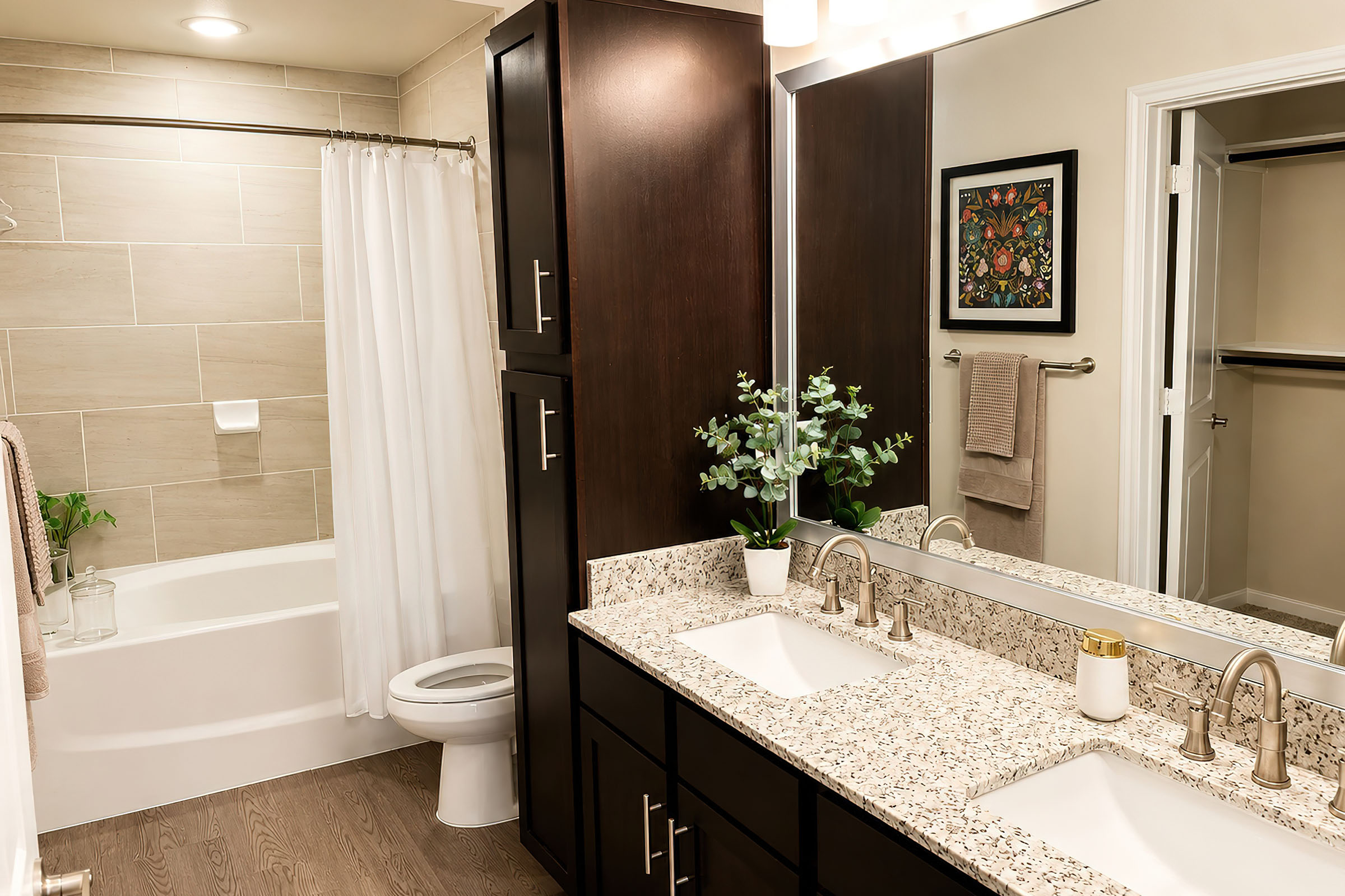 A modern bathroom featuring a double sink vanity with dark wood cabinets, a large mirror, and a decorative plant. The space includes a white bathtub with a shower curtain, light-colored tile walls, and wooden flooring. A towel rack with a beige towel is visible, along with artwork on the wall.