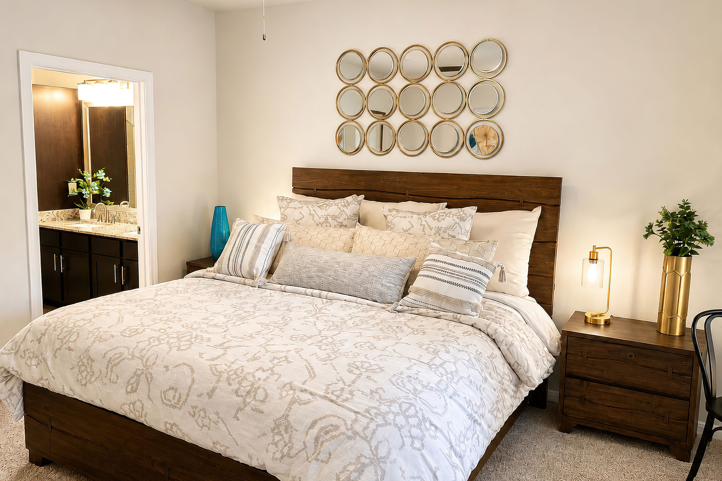 A cozy bedroom featuring a wooden bed with decorative pillows and a patterned blanket. There's a nightstand with a gold lamp and a small plant. Above the bed, a circular mirror arrangement adds visual interest. A doorway in the background leads to a bathroom area with dark wood cabinets. The room has light, neutral colors.