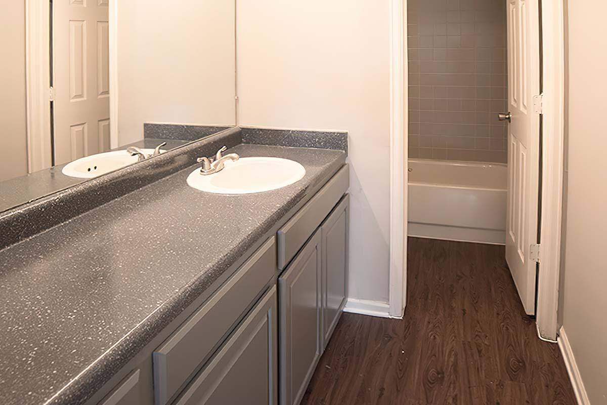 A clean bathroom featuring a gray countertop with double sinks, gray cabinets, and a large mirror. The space is well-lit with light walls and a wooden floor. A doorway in the background leads to a tub-shower combo area, which has light tiles. Overall, the design is simple and modern.
