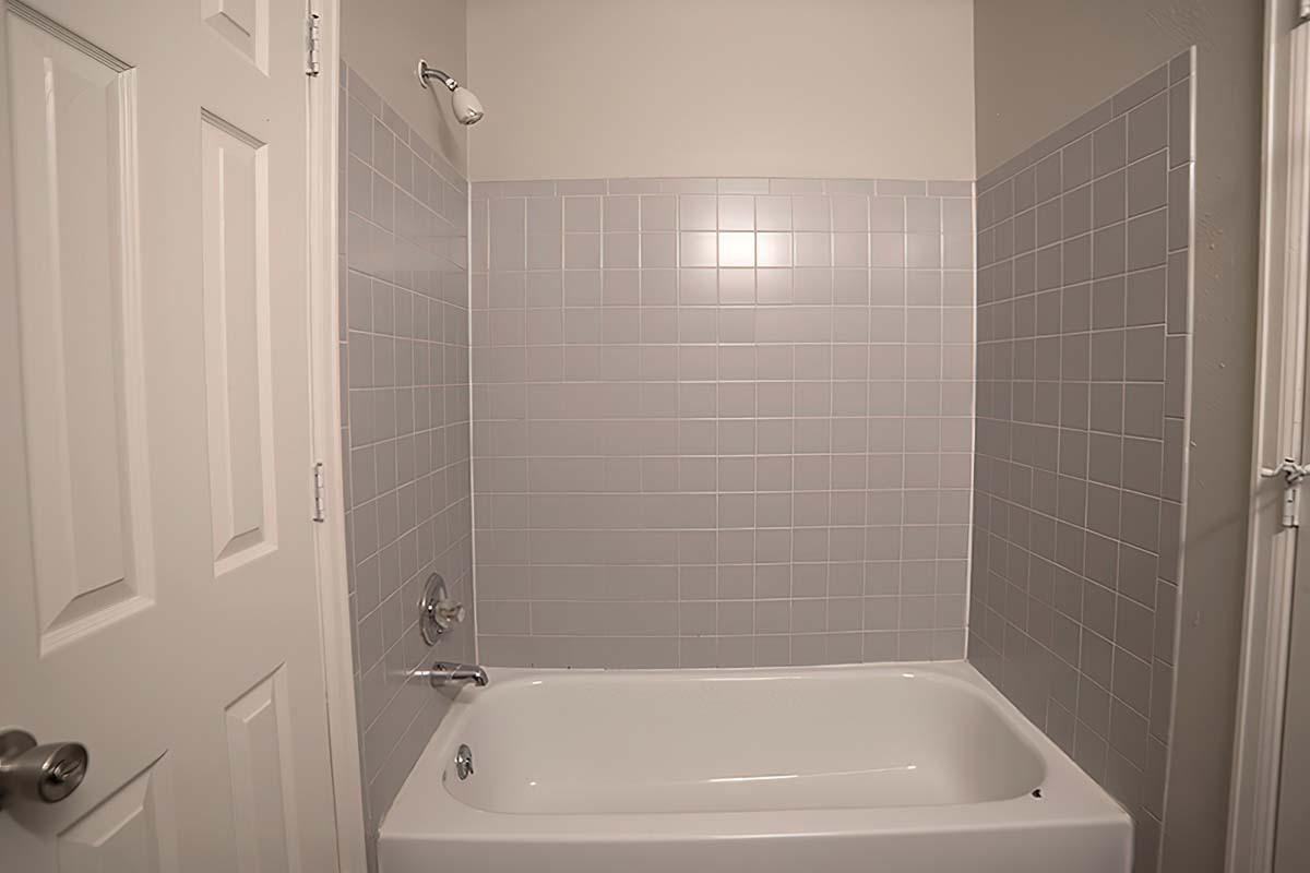 A clean, empty bathroom with a gray tiled shower area and a white bathtub. The walls are tiled halfway up, and there is a showerhead on the wall. A white door is partially visible, and the overall lighting is bright, creating an open and fresh feel.