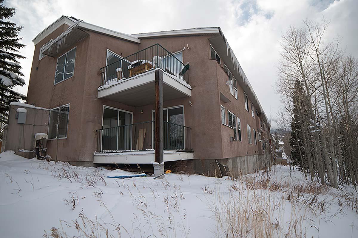 A two-story building situated in a snowy landscape, featuring multiple balconies and large windows. The exterior is a light brown color, and there are trees in the background, indicating a winter setting. Snow covers the ground, creating a serene, quiet atmosphere.