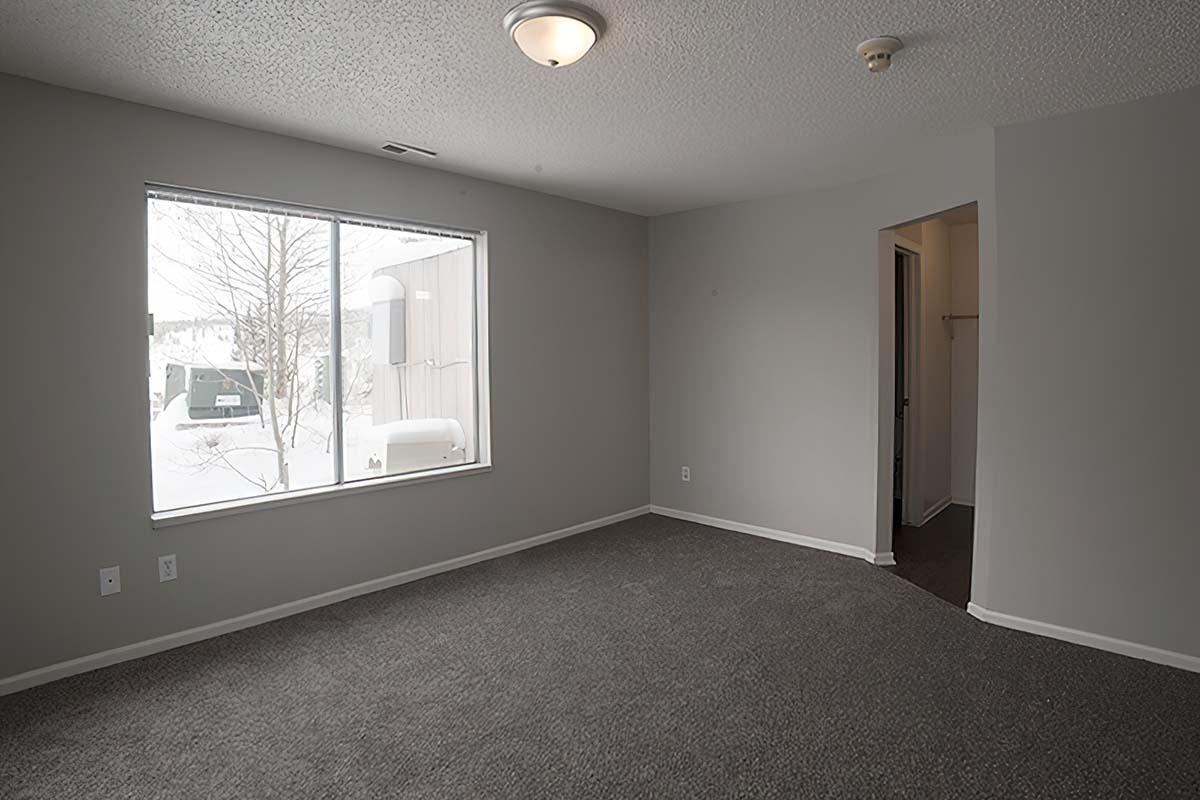A vacant room with gray walls and carpet, featuring a large window offering a view of a snowy landscape outside. The room has a ceiling light fixture and a doorway leading to another area.