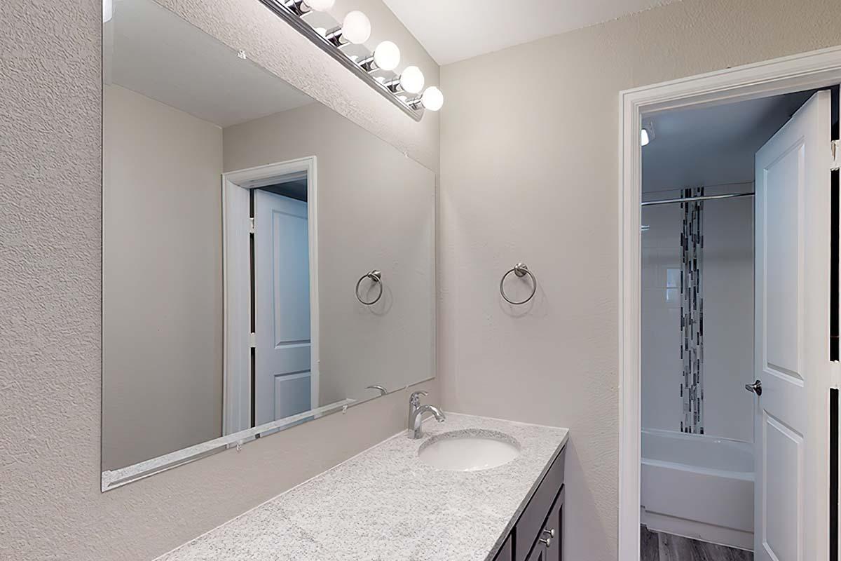 A modern bathroom featuring a double-sink vanity with a white granite countertop, a large rectangular mirror above the sinks, and a light fixture with multiple bulbs. The walls are painted in a neutral tone, and a door leads to a shower area visible through an open doorway.