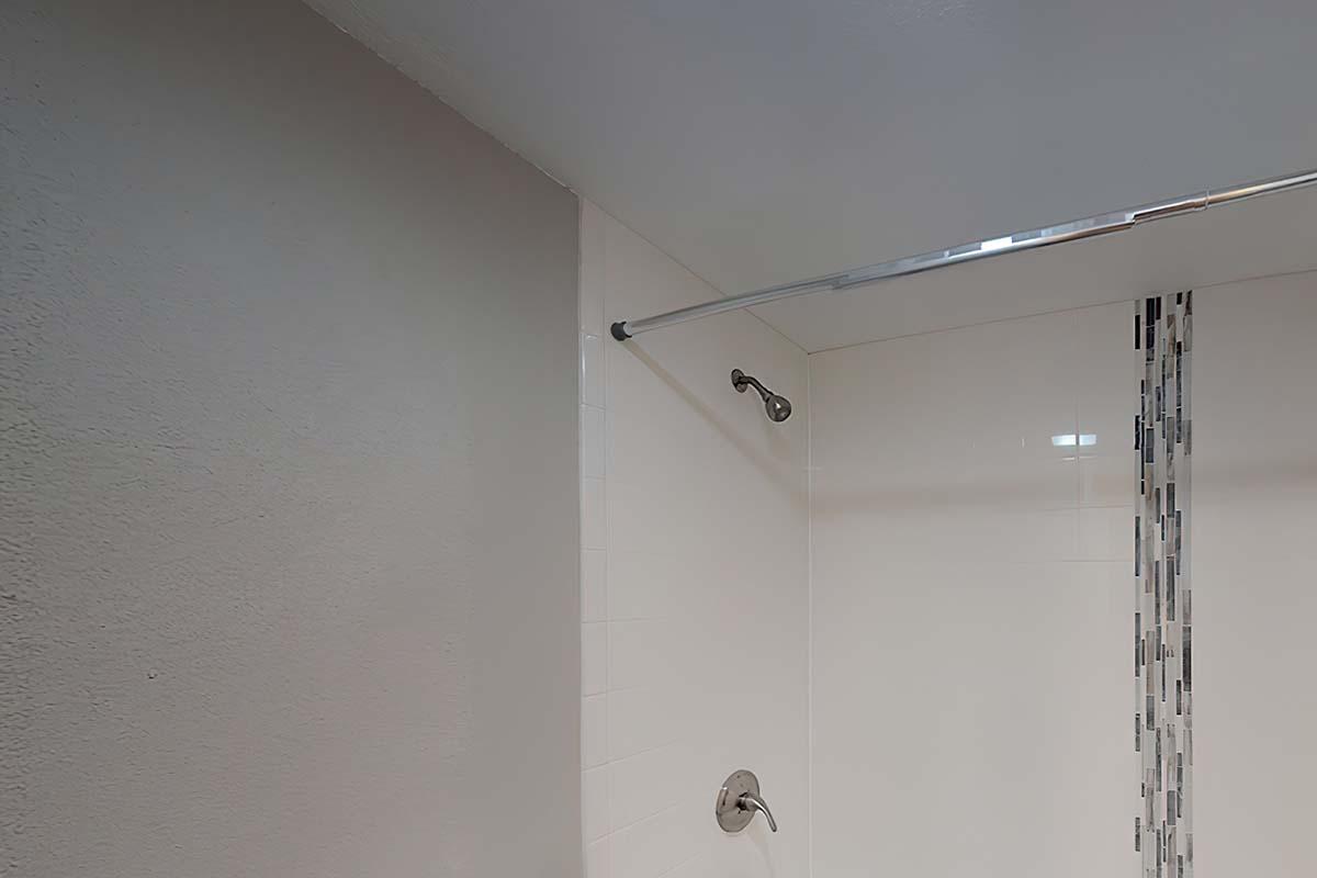 A clean bathroom shower area featuring white tiles with a decorative strip. The shower rod is installed on the wall, ready for a curtain, and the floor appears to be tiled. The overall design is modern and minimalist with neutral colors.