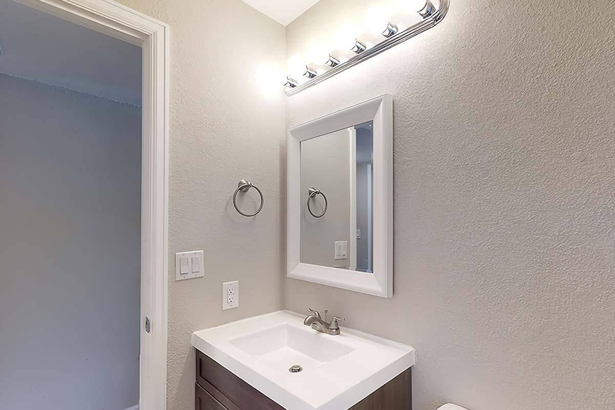 A modern bathroom featuring a vanity with a white sink and faucet, a large rectangular mirror, and a light fixture with multiple bulbs above. The walls are painted a light gray, and there is a towel bar on the wall beside the mirror. The door is slightly ajar, indicating access to another area.