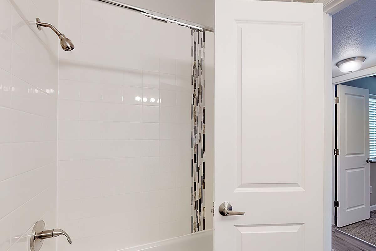 A bathroom featuring a white tiled shower area with a glass shower curtain, a silver showerhead, and a partially open door. A second door is visible in the background, leading to another room with soft lighting. The overall atmosphere is clean and modern.
