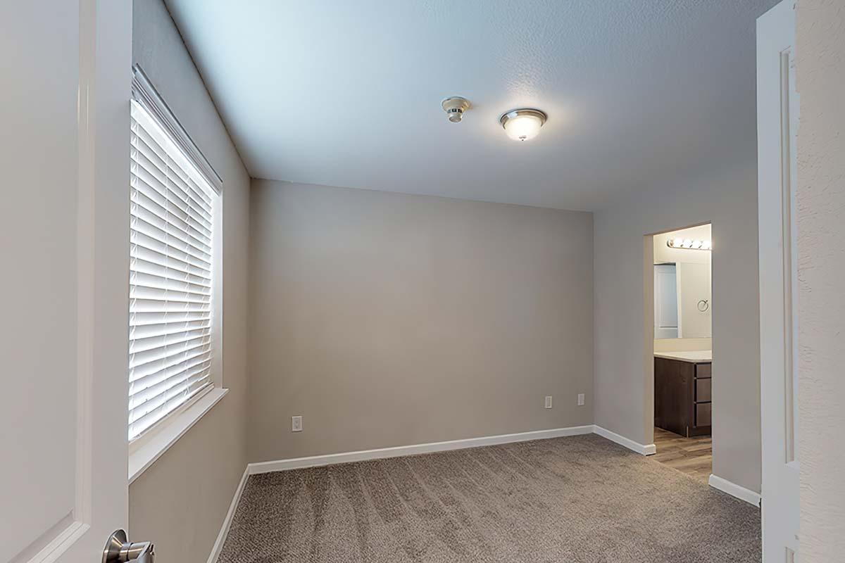 A well-lit, empty room with light gray walls and plush carpet. A window with white blinds allows natural light, and there's a ceiling light fixture. To the right, a doorway leads to a bathroom with a sink and mirror visible. The overall atmosphere is clean and inviting.
