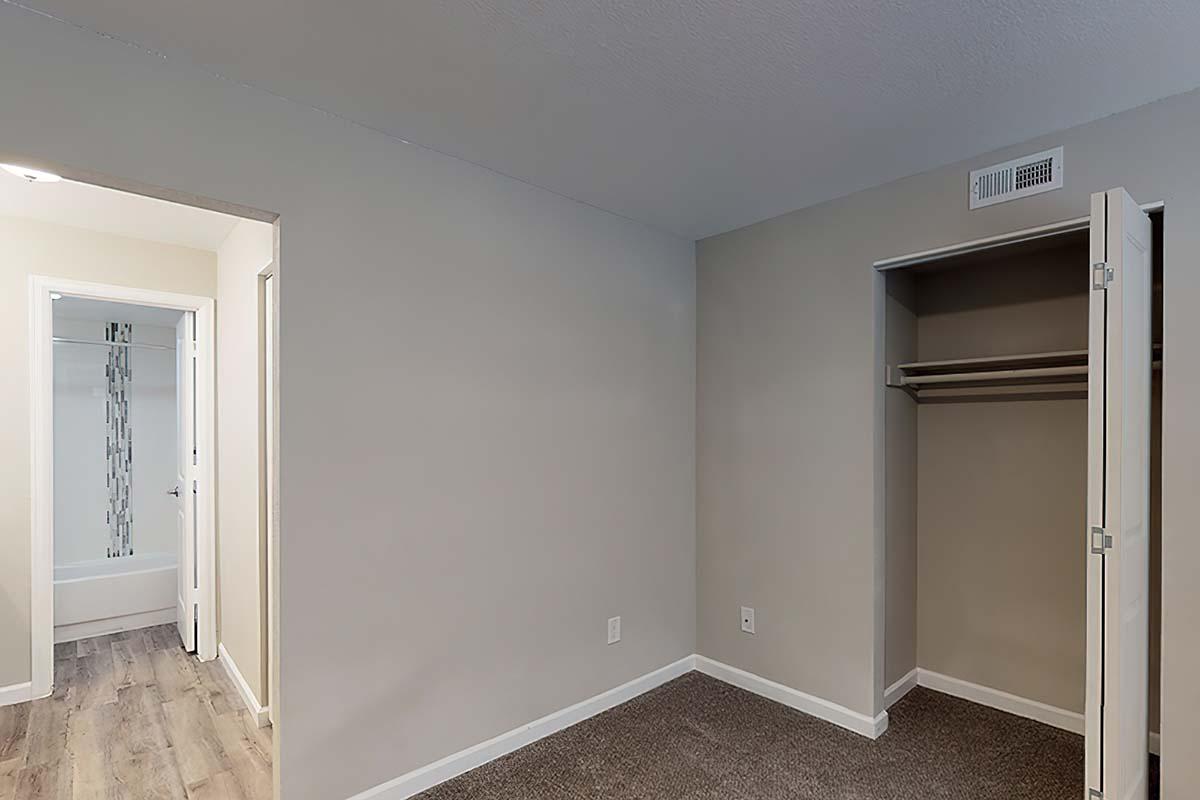 A small, empty bedroom featuring light gray walls and a closet with sliding doors. The floor is carpeted, and an open doorway leads to a bathroom with a visible bathtub. Natural light enters from the hallway.