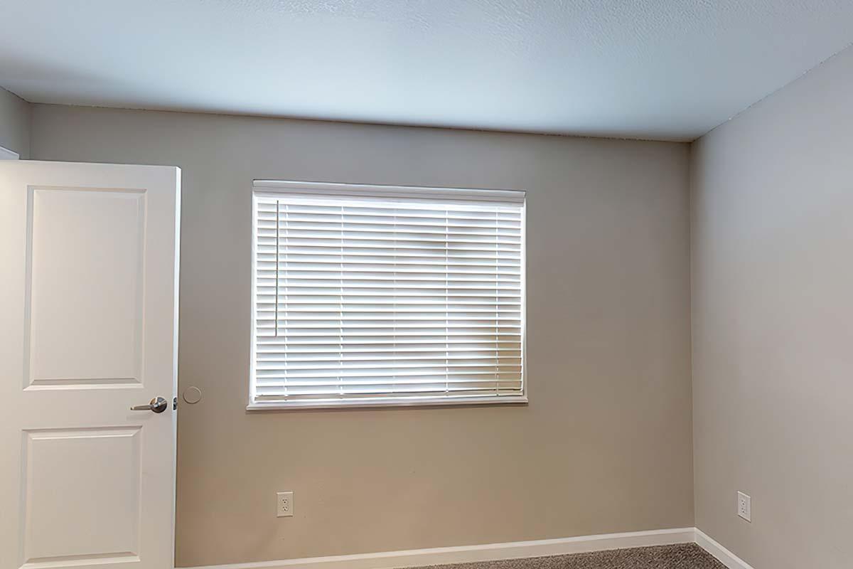 A light-colored room featuring a closed door on the left and a window on the right with horizontal blinds. The walls are painted in a neutral tone, and the floor is covered with carpeting. The overall appearance is clean and simple, lacking furniture or decor.