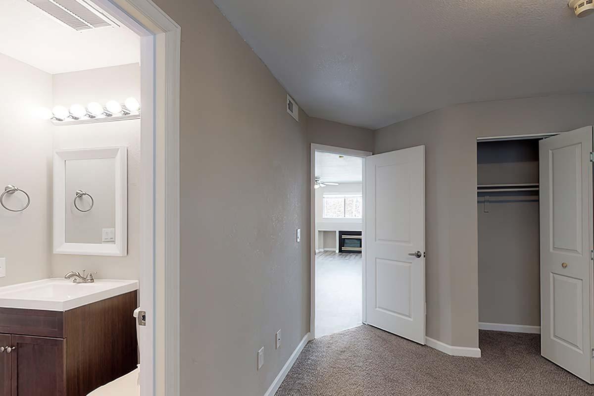 Interior view of a room showing a doorway leading to another space, a closet on the right, and a bathroom area with a vanity and mirror visible on the left. The walls are painted in a neutral tone, and the flooring appears to be carpeted. Natural light is coming from the entrance to the other room.