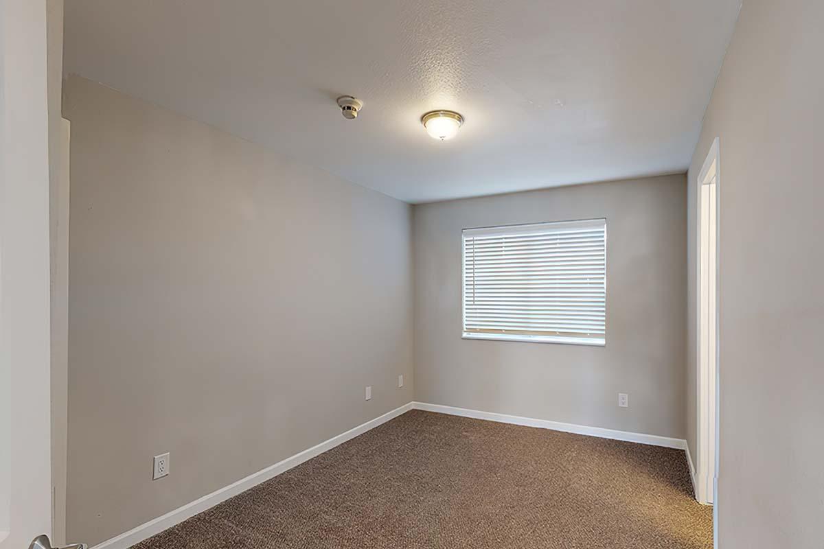 A vacant room with gray walls and beige carpet. There is a single window with white blinds, a ceiling light fixture, and a door on the left leading outside the frame. The space is empty and well-lit, creating a simple, minimalistic atmosphere.