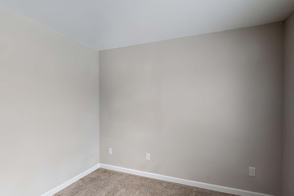 Empty room with light beige walls and a carpeted floor. The wall has two electrical outlets, and there is no furniture or decor present, giving it a clean and minimalistic appearance. The ceiling is white, and the overall space feels open and bright.