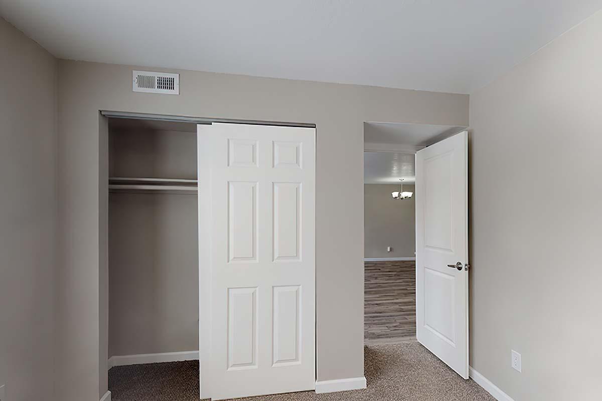 A light gray room featuring an open closet with sliding doors on the left and an adjoining door to another room on the right. The walls are painted in a neutral tone, and the floor has a carpeted texture. Natural light illuminates the spacious interior.