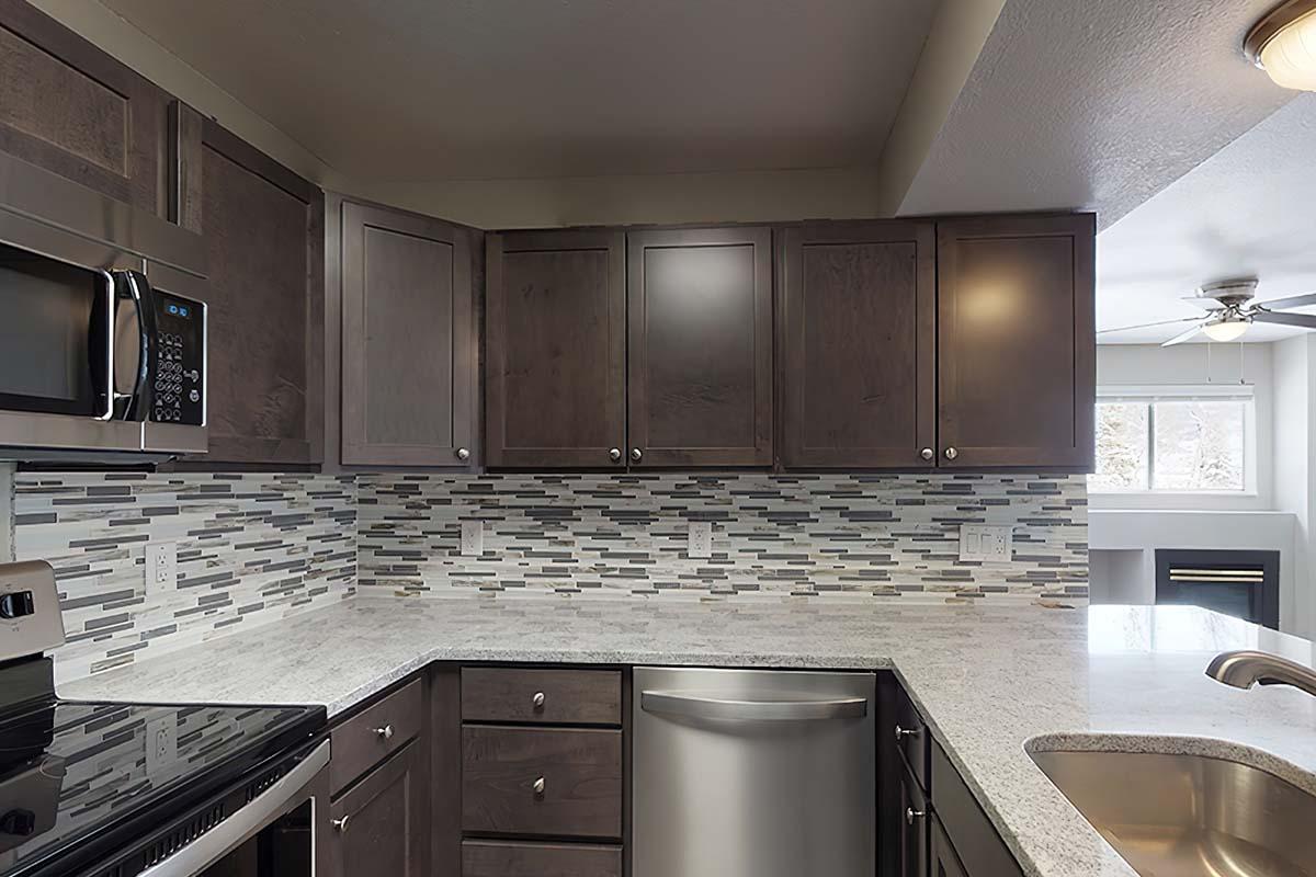 Modern kitchen featuring dark wood cabinets, a stainless steel refrigerator, and an electric stove. The countertop is gray with a textured finish, and a stylish backsplash made of glass tiles adorns the wall. Natural light streams in from a window, creating a bright and inviting atmosphere.
