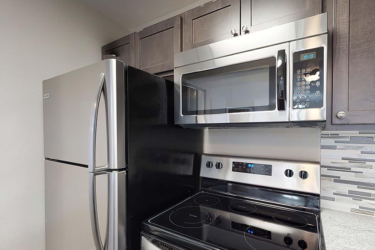 A modern kitchen with stainless steel appliances, including a refrigerator, microwave, and an electric range. The cabinetry above is dark wood, and the countertop features a light-colored surface. The setting has a contemporary design.