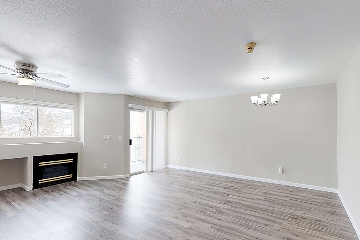 Spacious, empty living room featuring light wood flooring, a ceiling fan, a white fireplace, and large windows providing natural light. The room has neutral-colored walls and a sliding glass door leading to a balcony, complemented by a simple chandelier overhead.